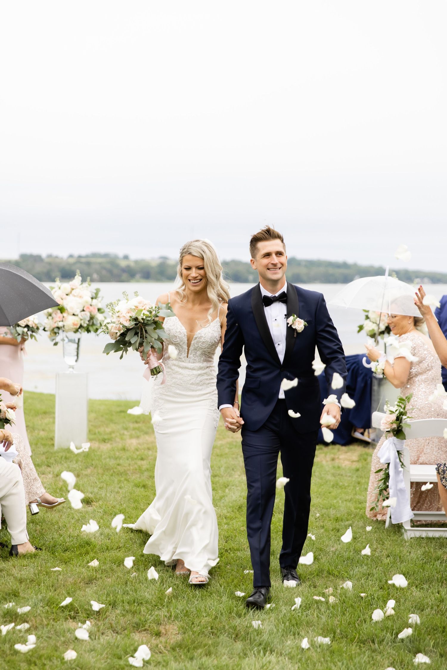 Newlyweds walk down aisle amid flower petals at lakeside outdoor ceremony.