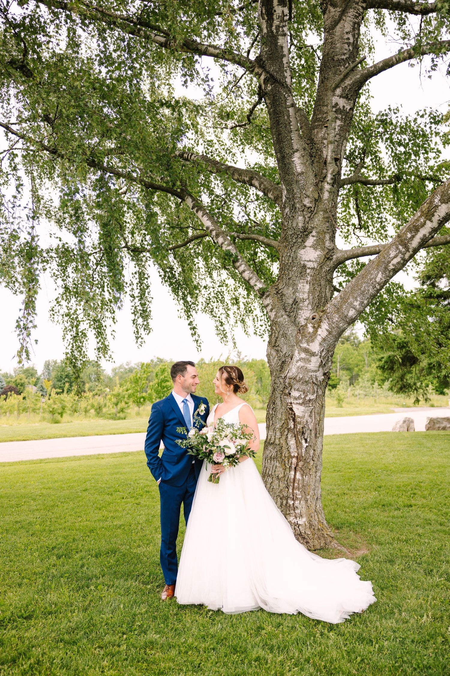 Wedding couple poses beneath majestic weeping willow tree on grassy lawn.