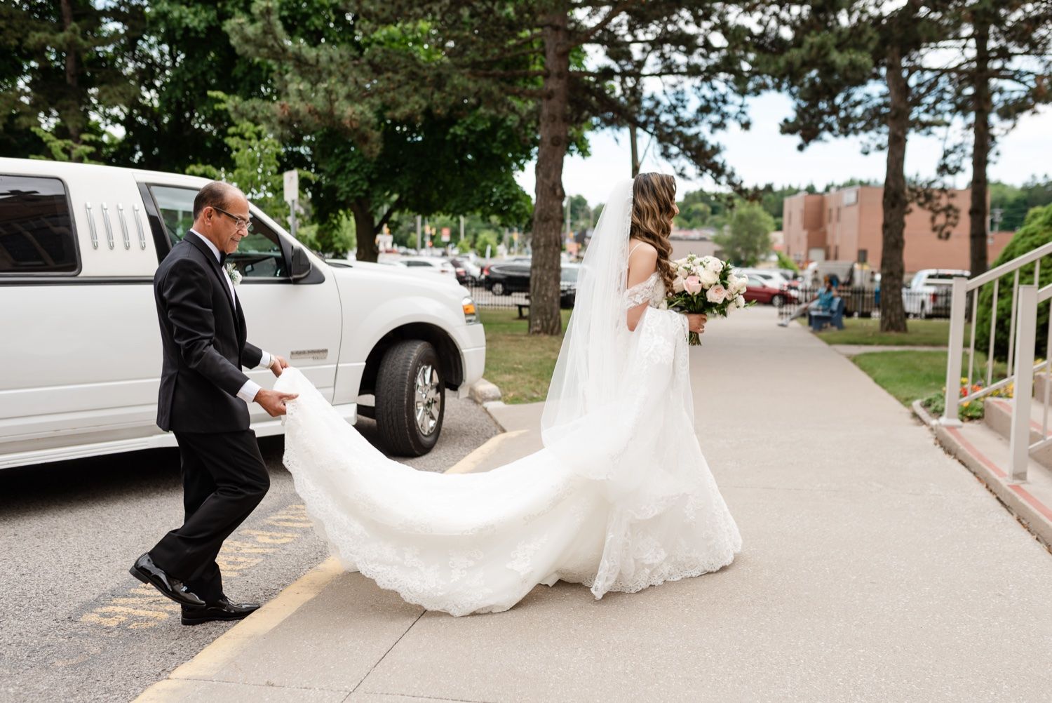 A wedding couple walking outdoors with flowing white veil trailing behind near limousine.