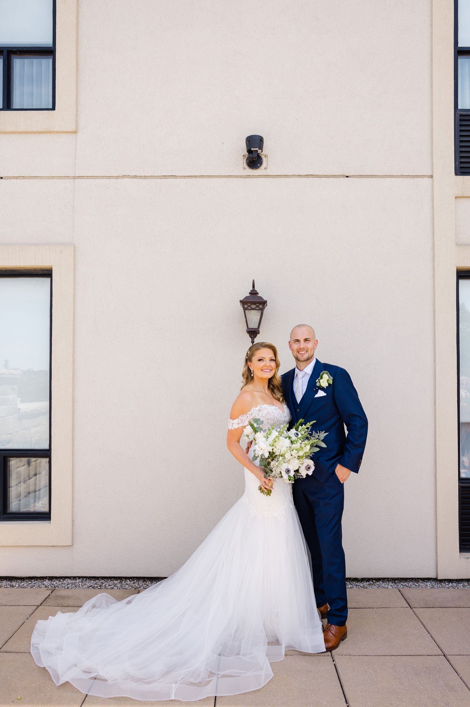 Elegant wedding portrait against beige wall with vintage lantern featuring couple in navy and white attire.