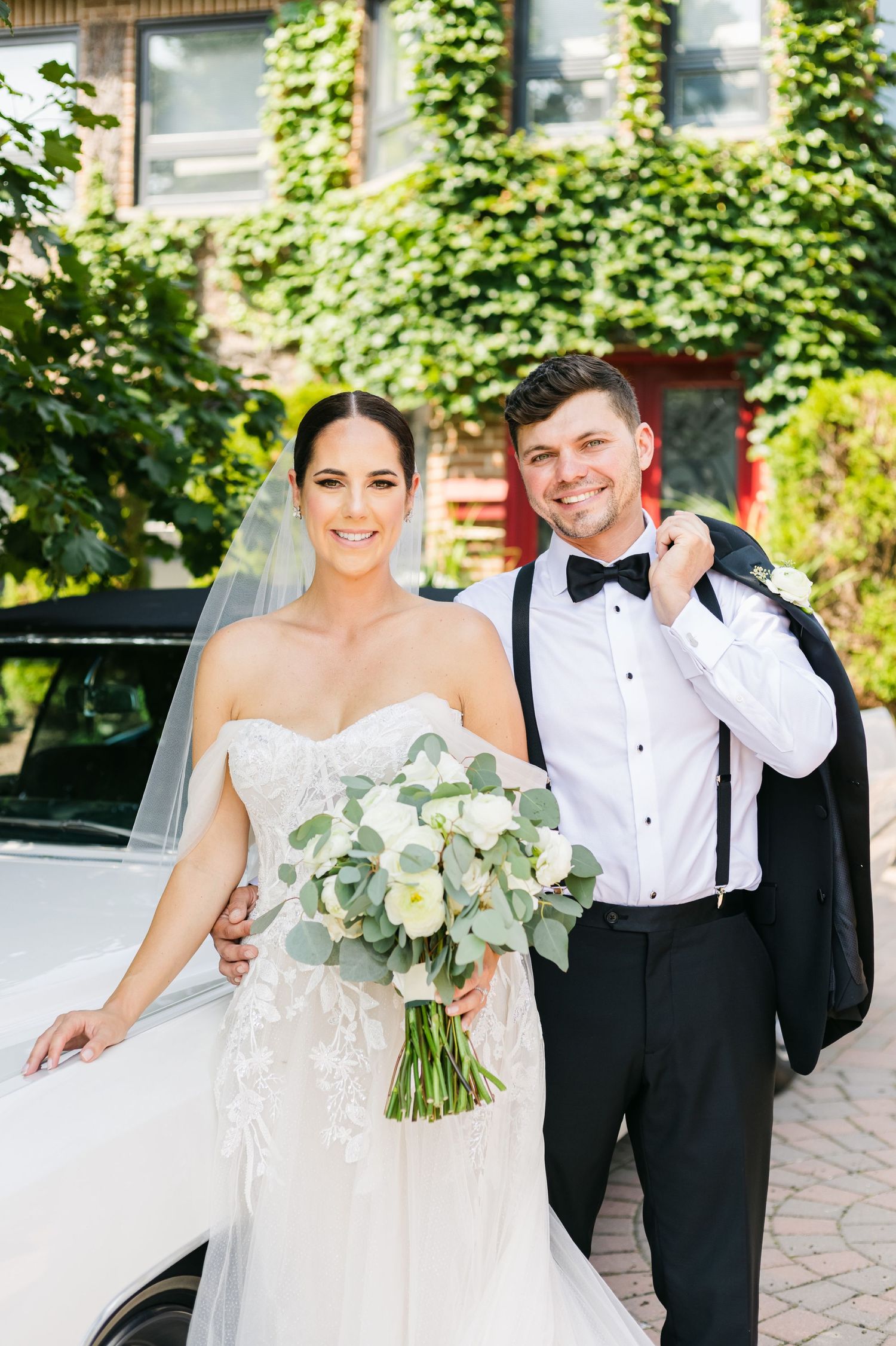 Wedding photo showcasing a classic white car and formal attire against an ivy-covered building backdrop.