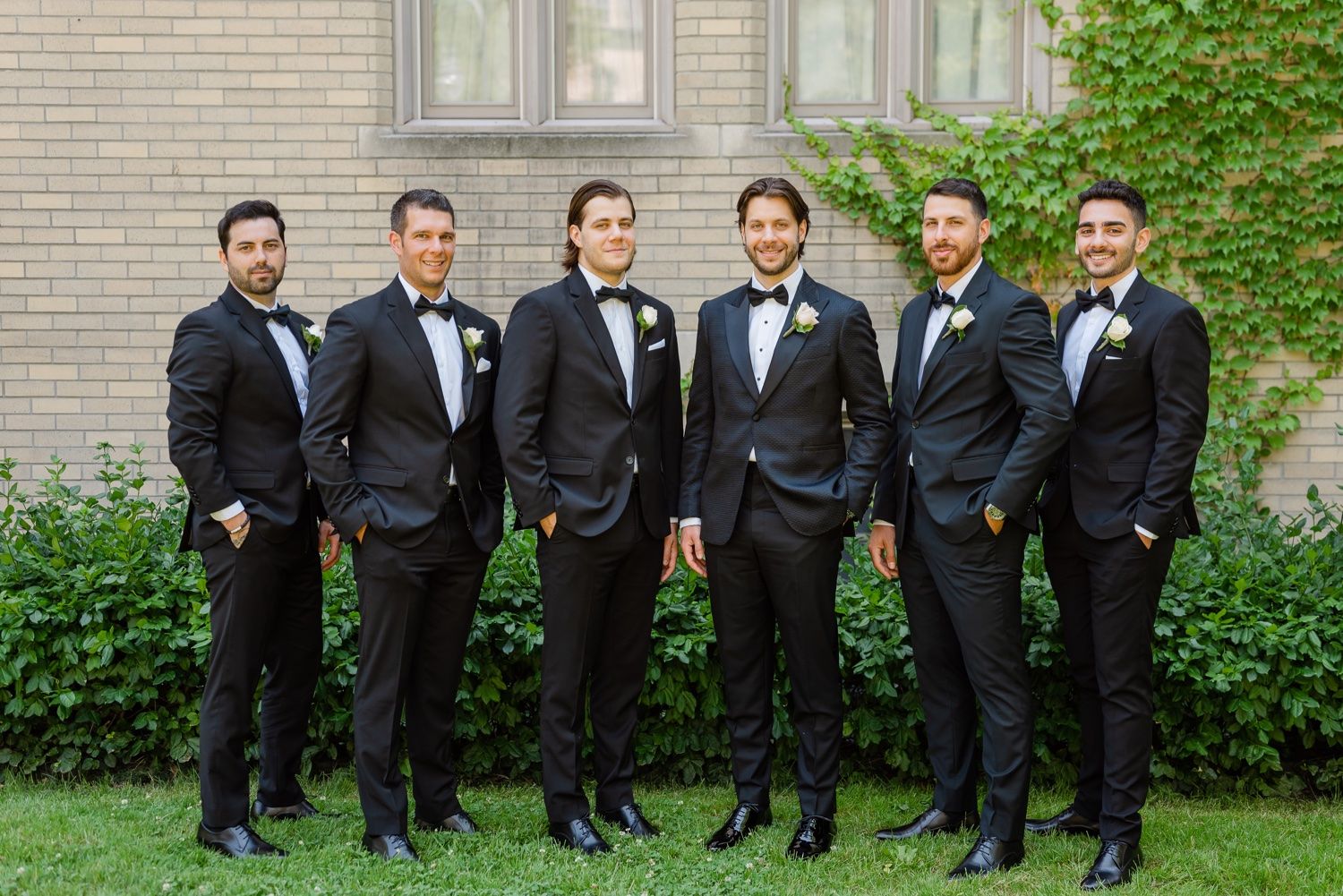Group of six groomsmen in matching black tuxedos stand against brick wall.