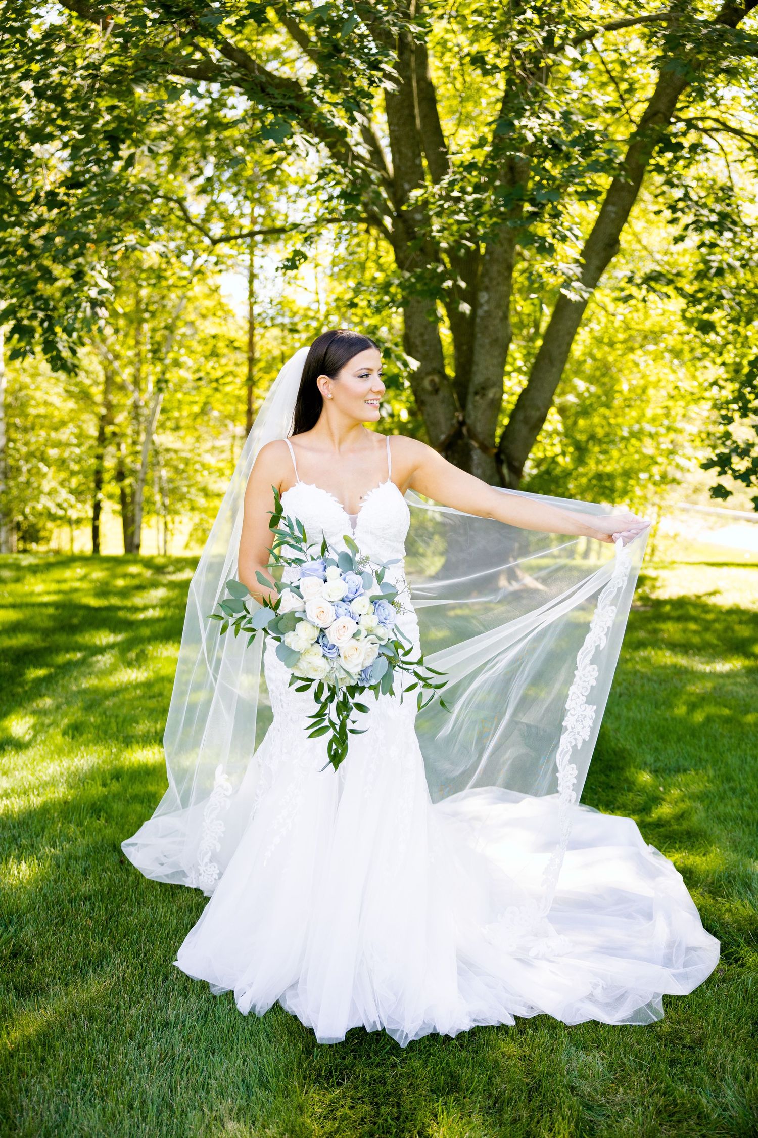 Bride in white gown poses with flowing veil in sunlit garden setting.