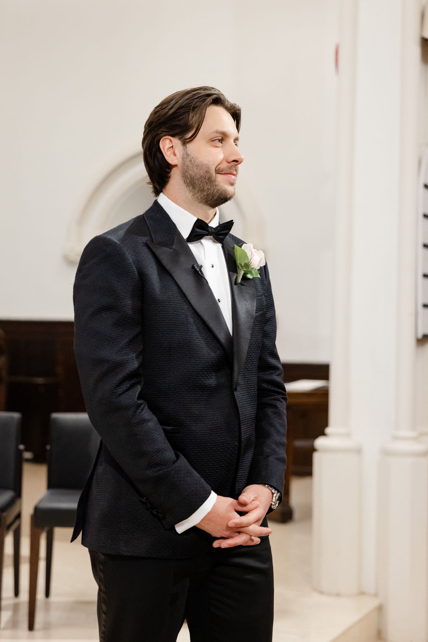 A groom in a black tuxedo and bowtie with a boutonniere smiling while standing indoors.