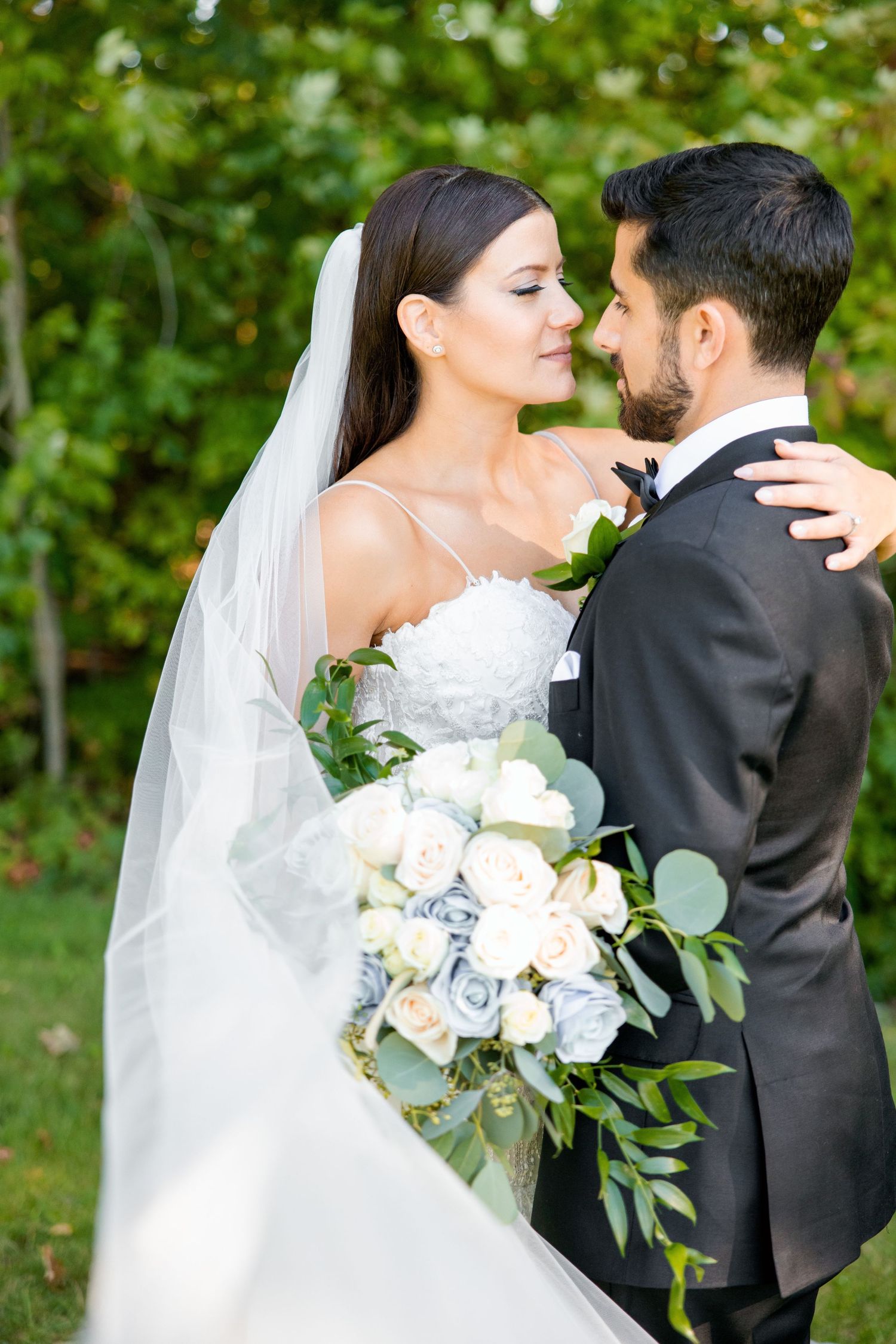Elegant outdoor wedding portrait with white floral bouquet and flowing veil against green foliage.