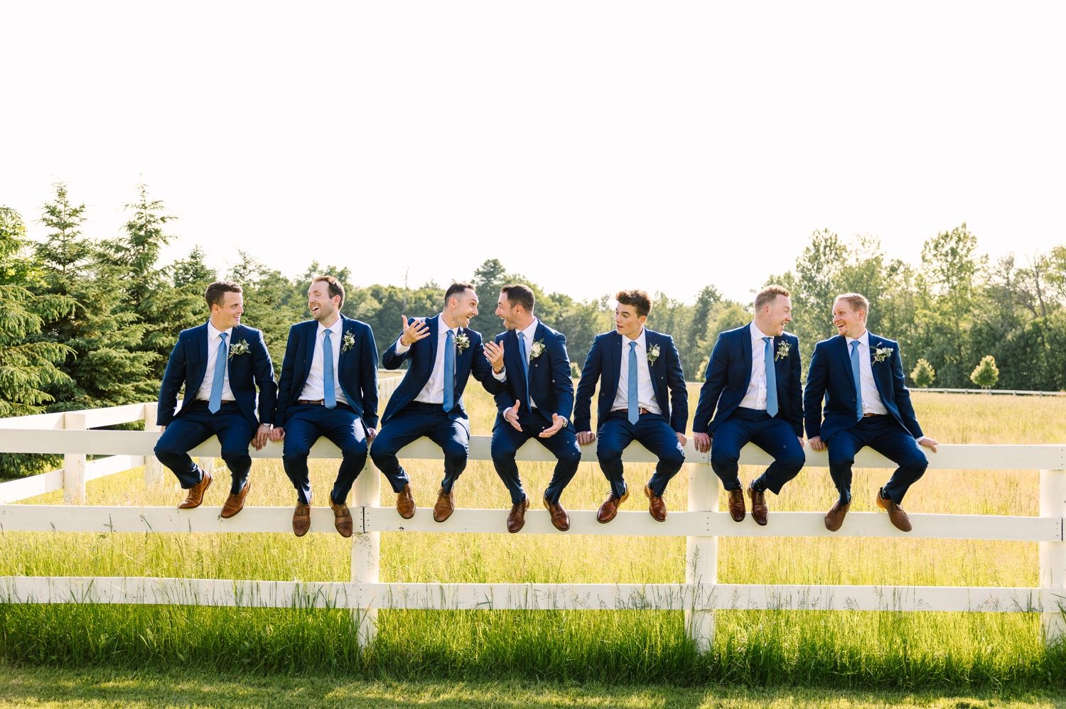 Group of groomsmen in navy suits sitting casually on white fence during wedding day.