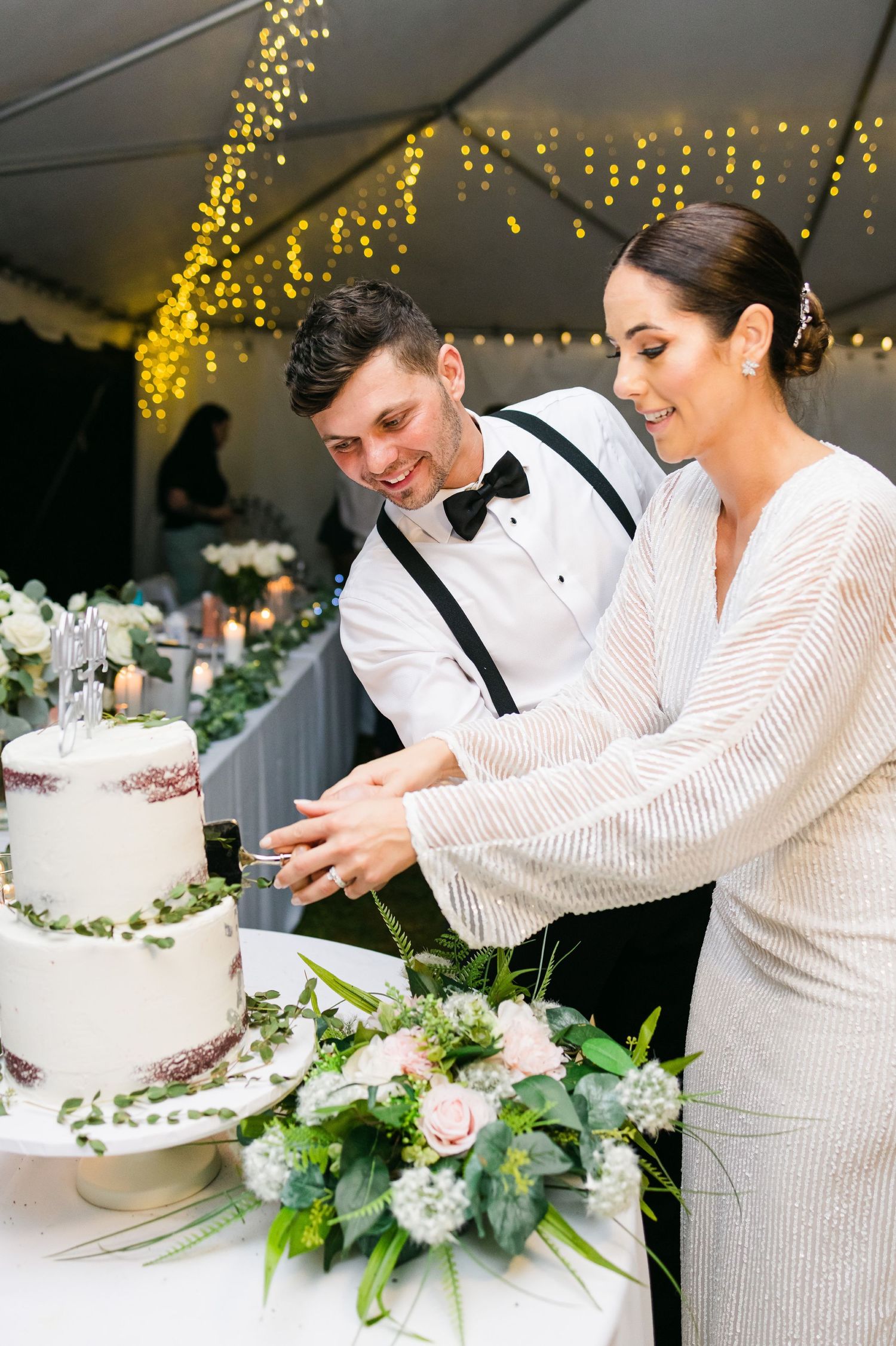 Couple cutting their white wedding cake decorated with fresh flowers and greenery.