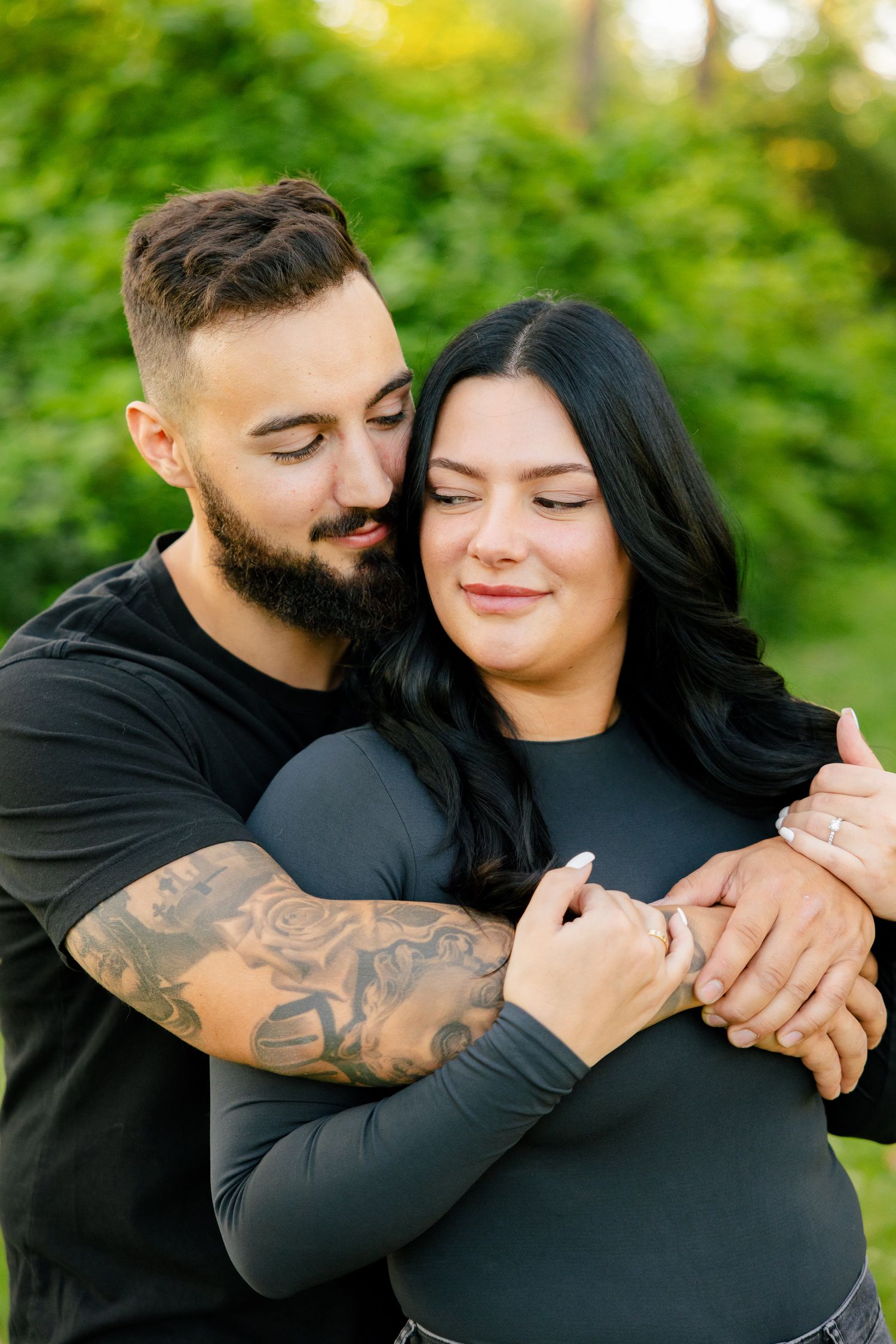 Couple shares intimate embrace in garden setting with man's tattooed arm holding partner from behind.
