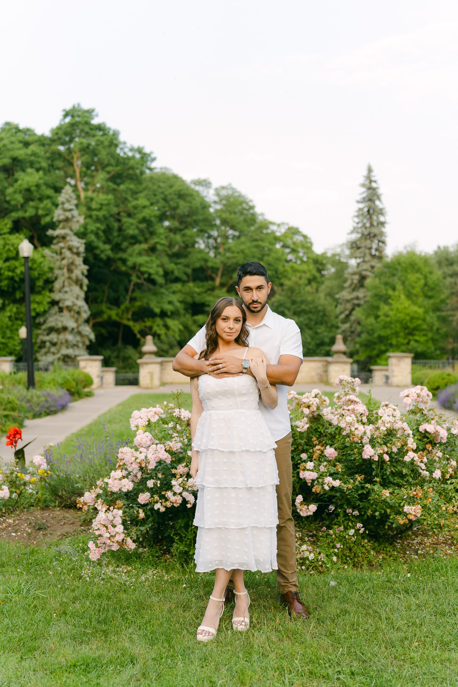 A full-length engagement portrait in a landscaped garden with stone features and roses.