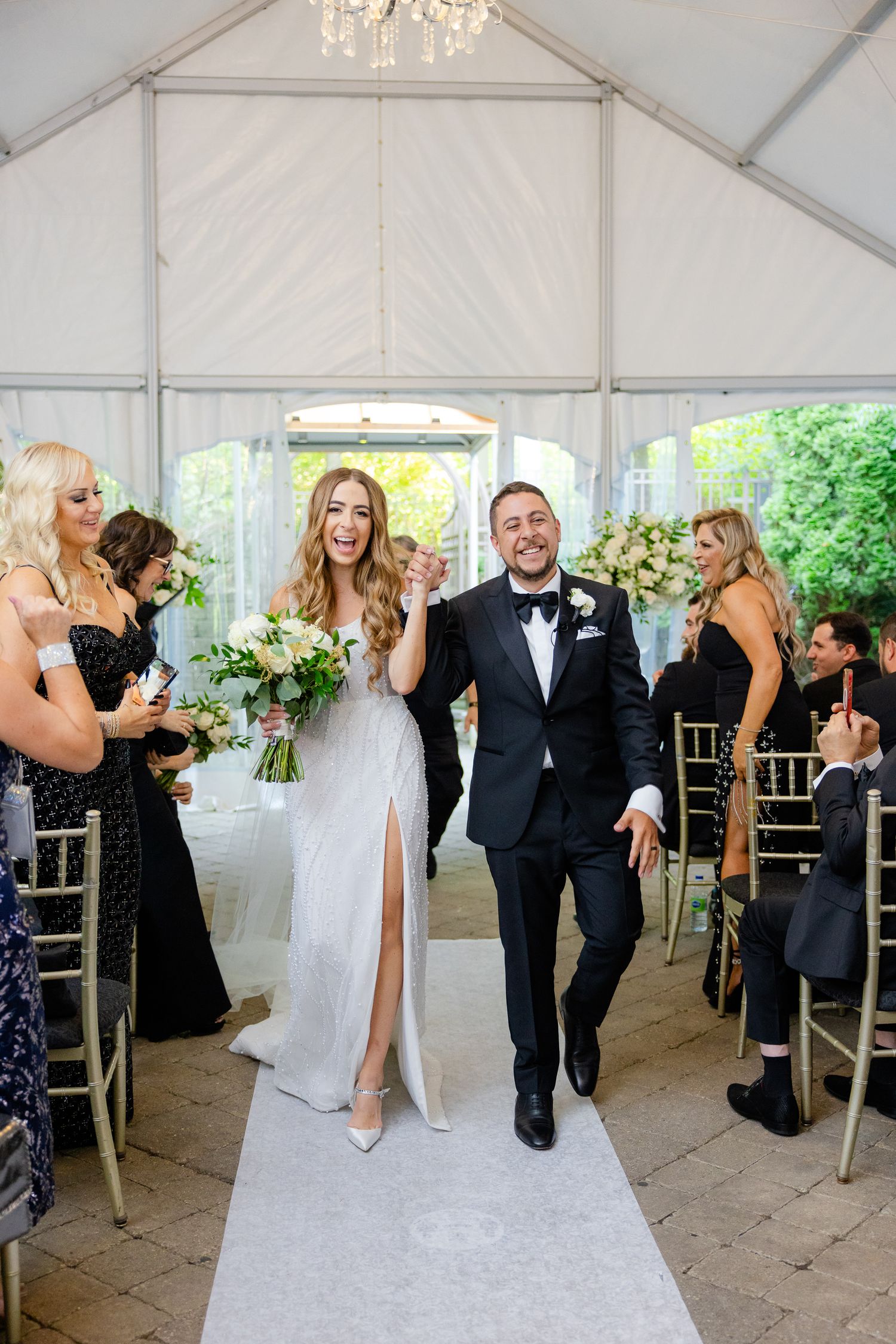 A couple makes their grand exit down a white aisle runner in a tent venue with guests looking on from gold chairs.