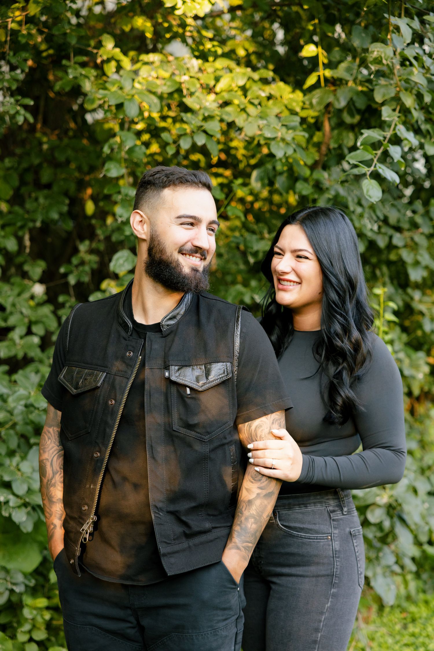 A couple smiles while posing together in front of green leaves and sunlit trees.