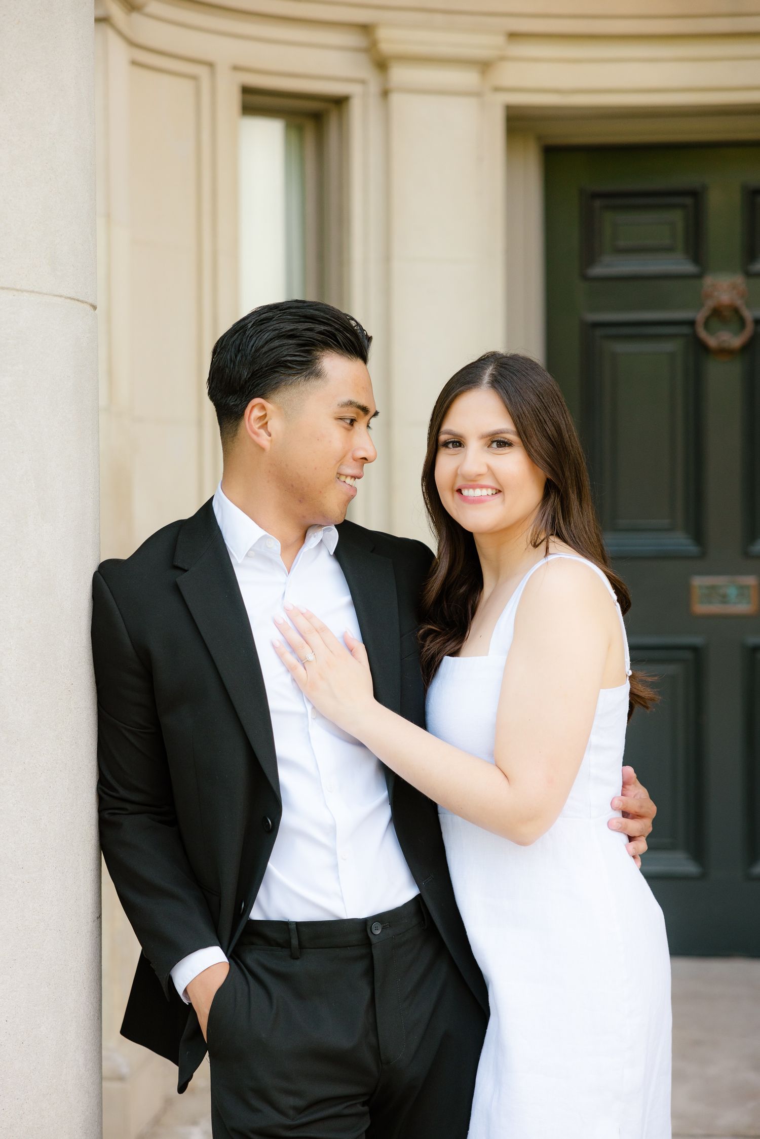 Couple in formal attire share affectionate moment against elegant architectural backdrop.