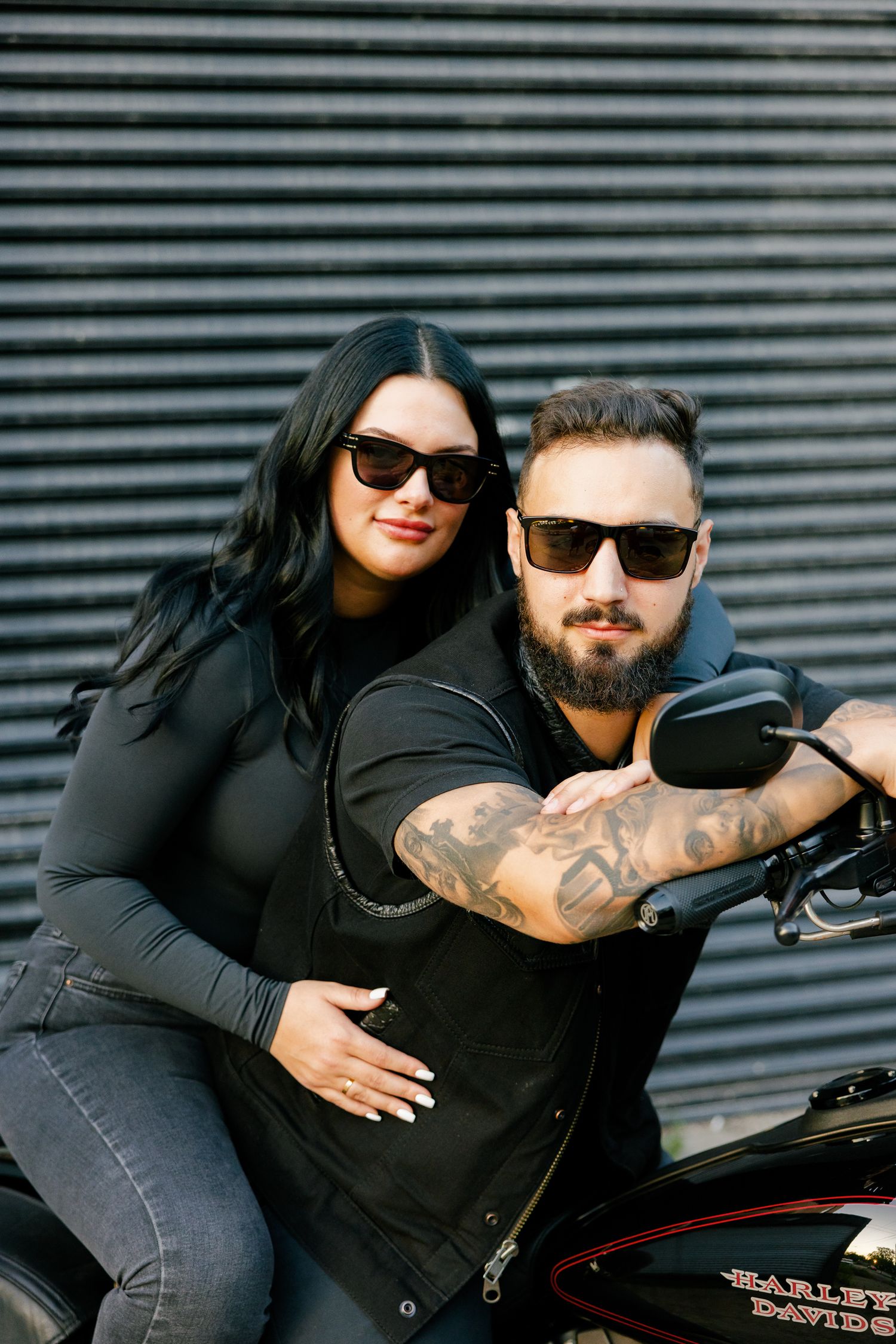 Couple in matching black outfits embraces on motorcycle against metal wall.