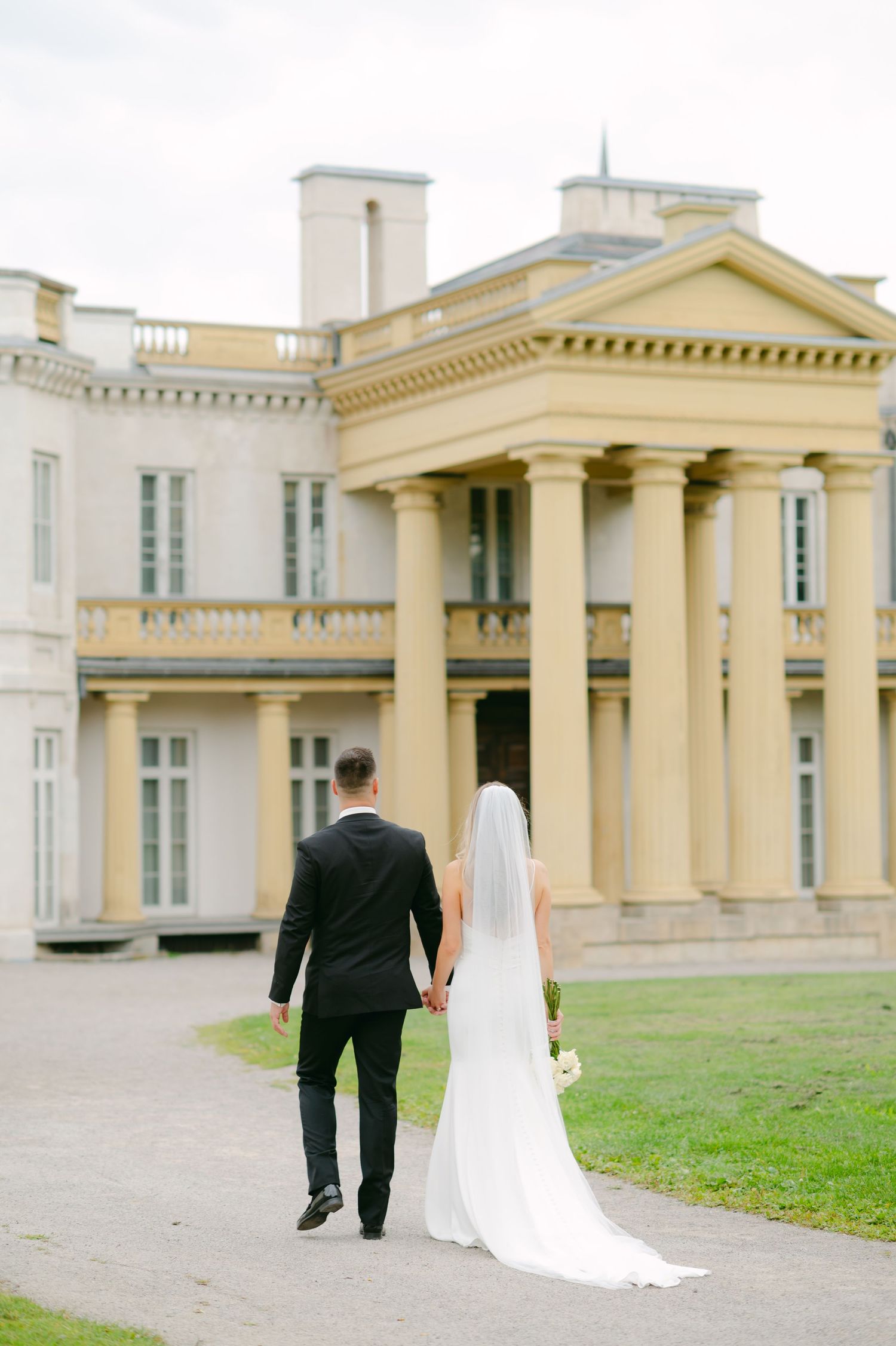 A wedding couple walking hand in hand towards an elegant yellow mansion with white columns.
