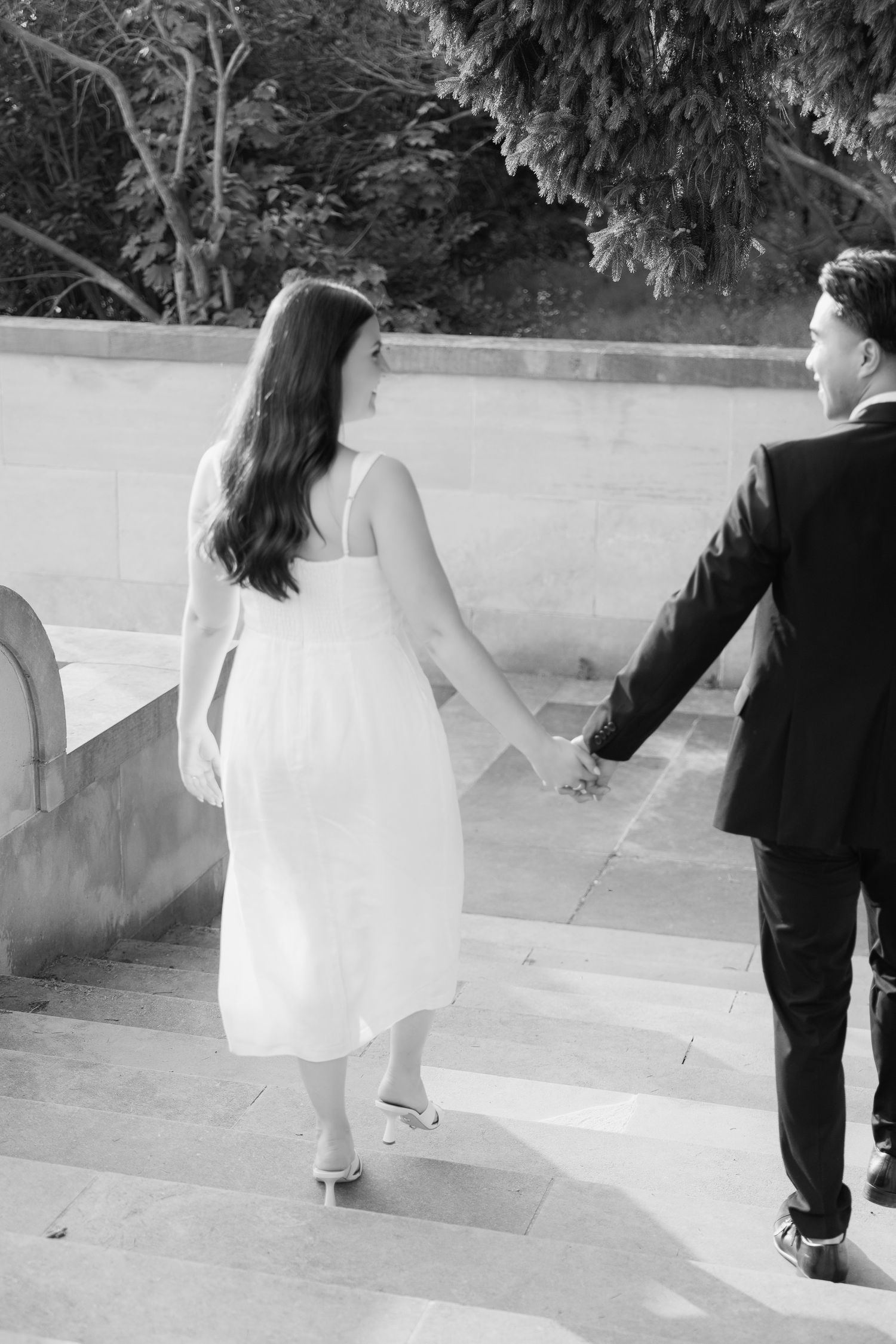 A black and white photo of a couple walking up stone steps hand in hand in formal attire.