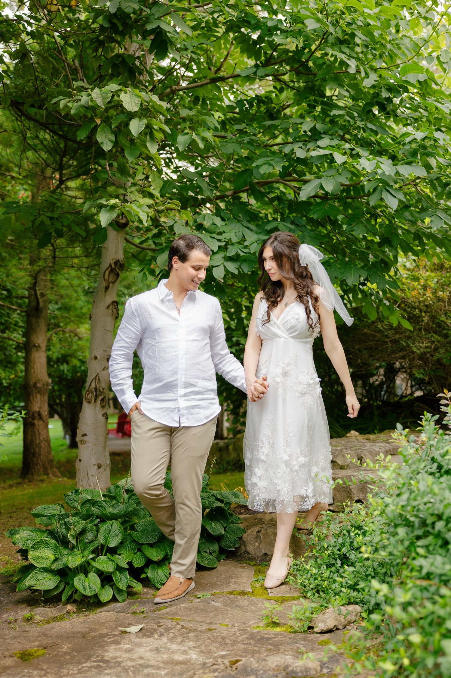 A couple takes a romantic stroll through a garden path under leafy green trees.