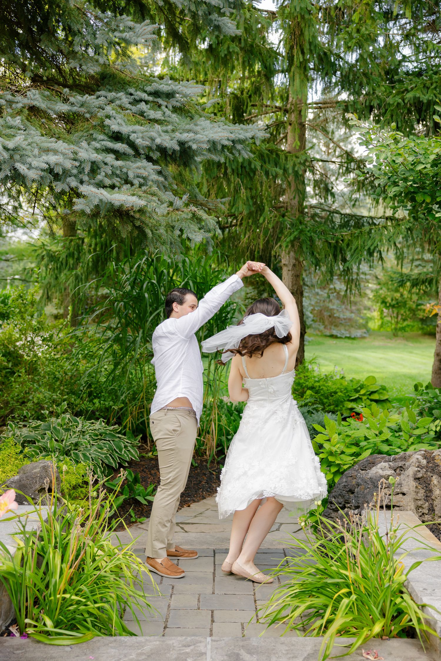 Dancing couple in wedding attire twirls on stone pathway surrounded by garden greenery.
