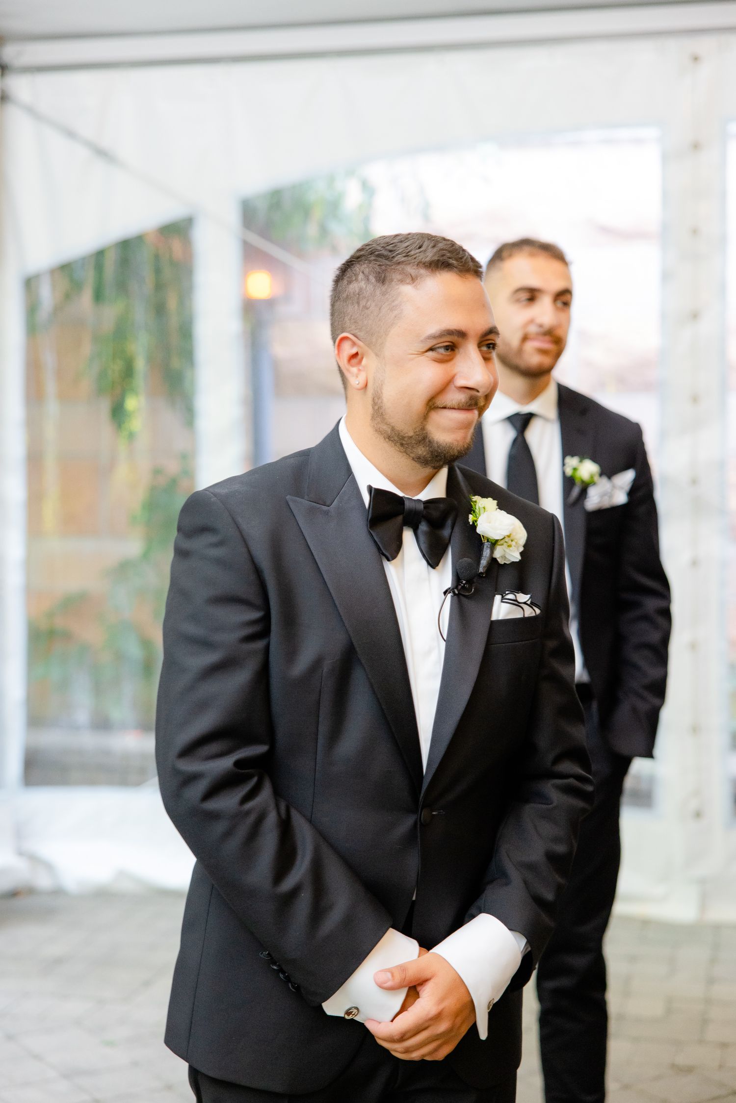 Groom and groomsman stand together in black tuxedos during formal wedding event.