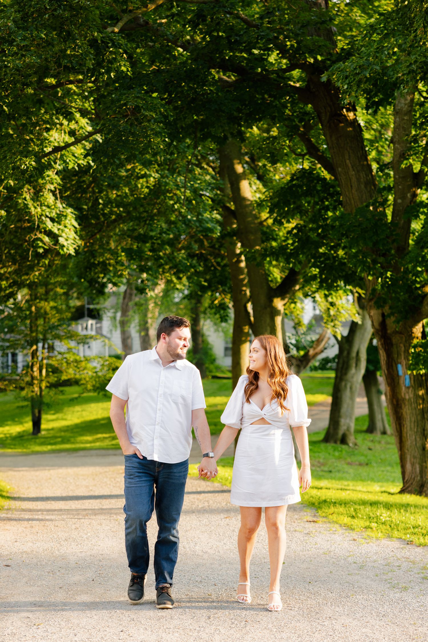 A couple holding hands strolls down a sunlit path surrounded by lush green trees.