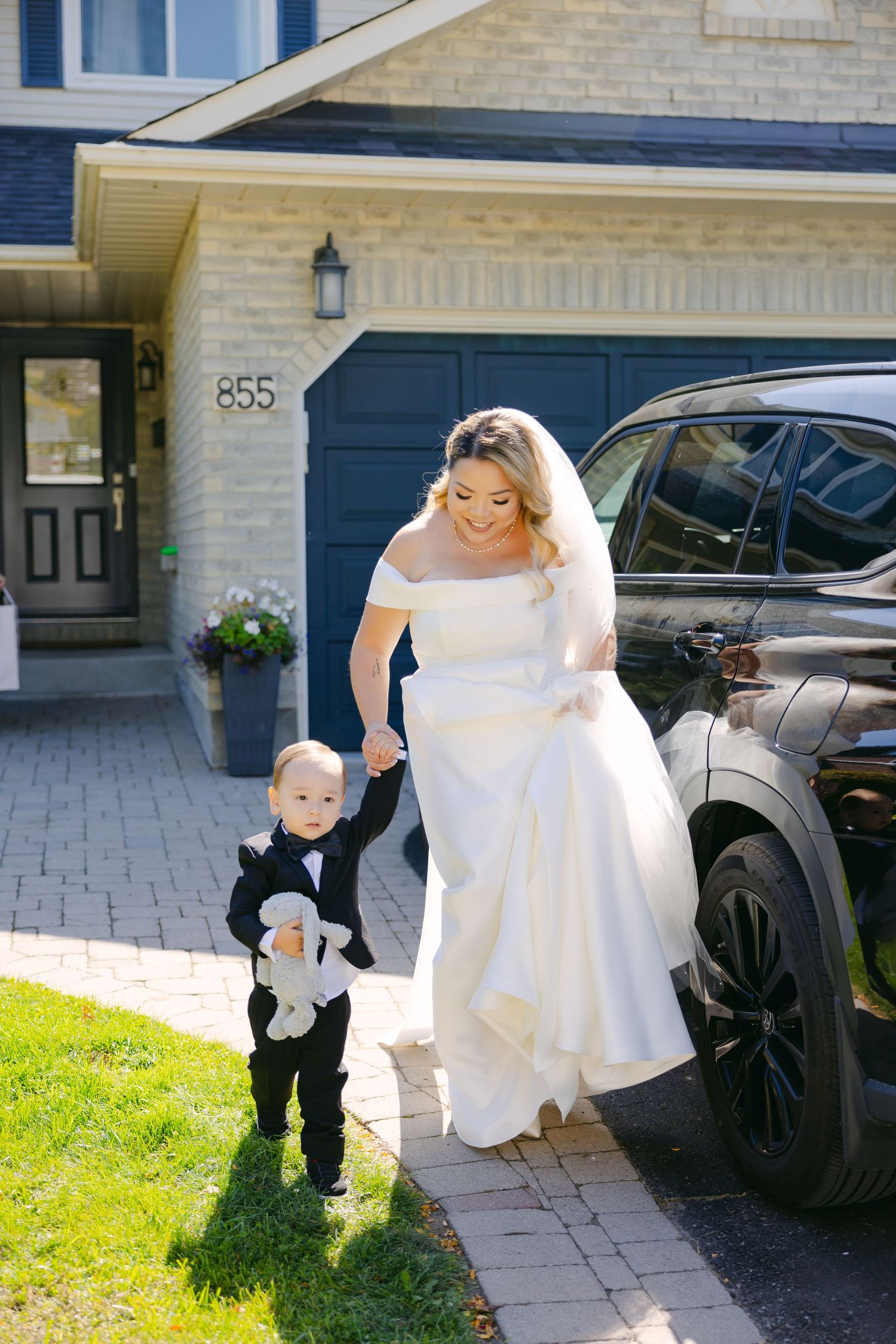 A bride in white helps a small child walk outside a house with a black car in the background.