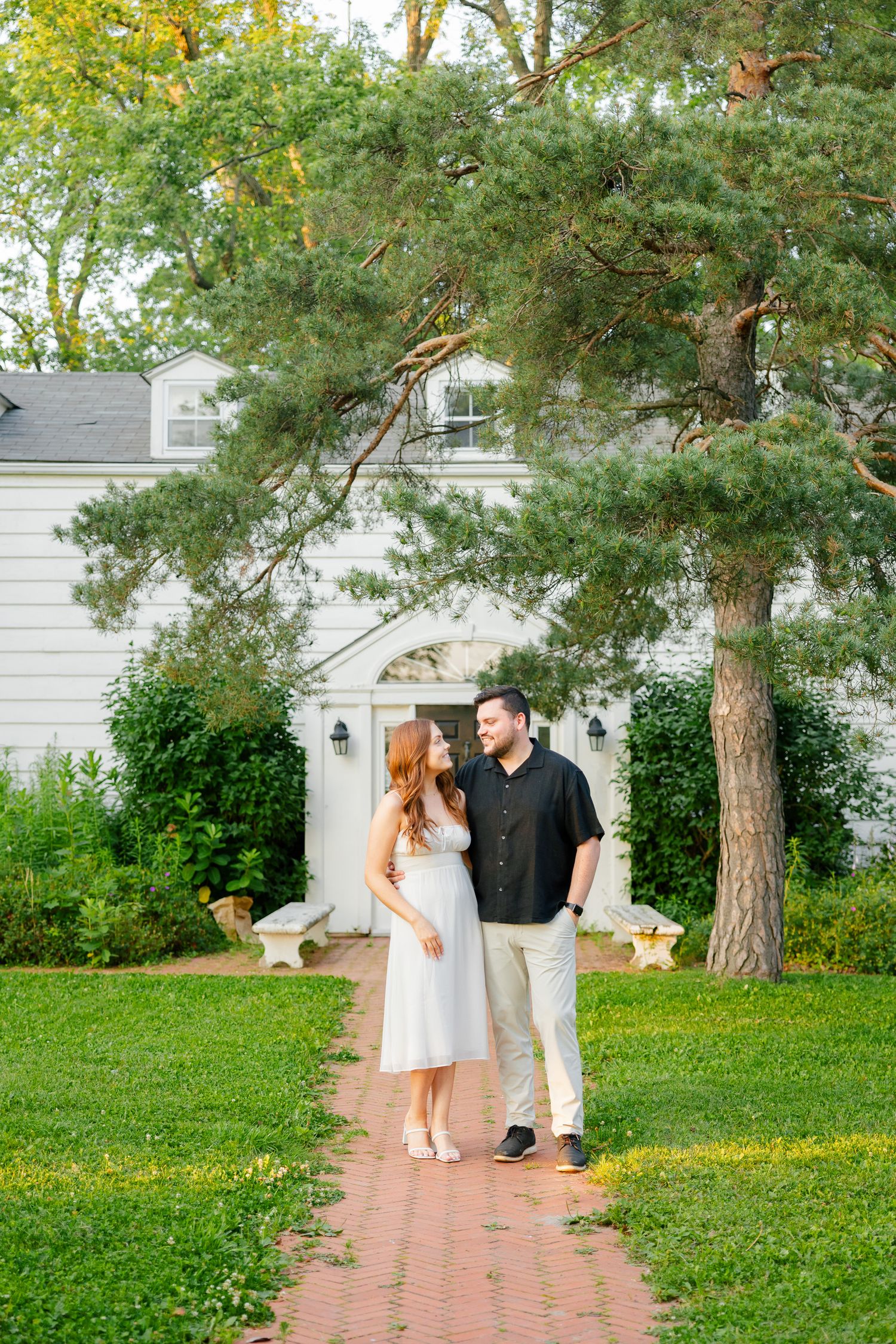 Two people share a tender moment on a brick walkway surrounded by lush greenery and a white house.