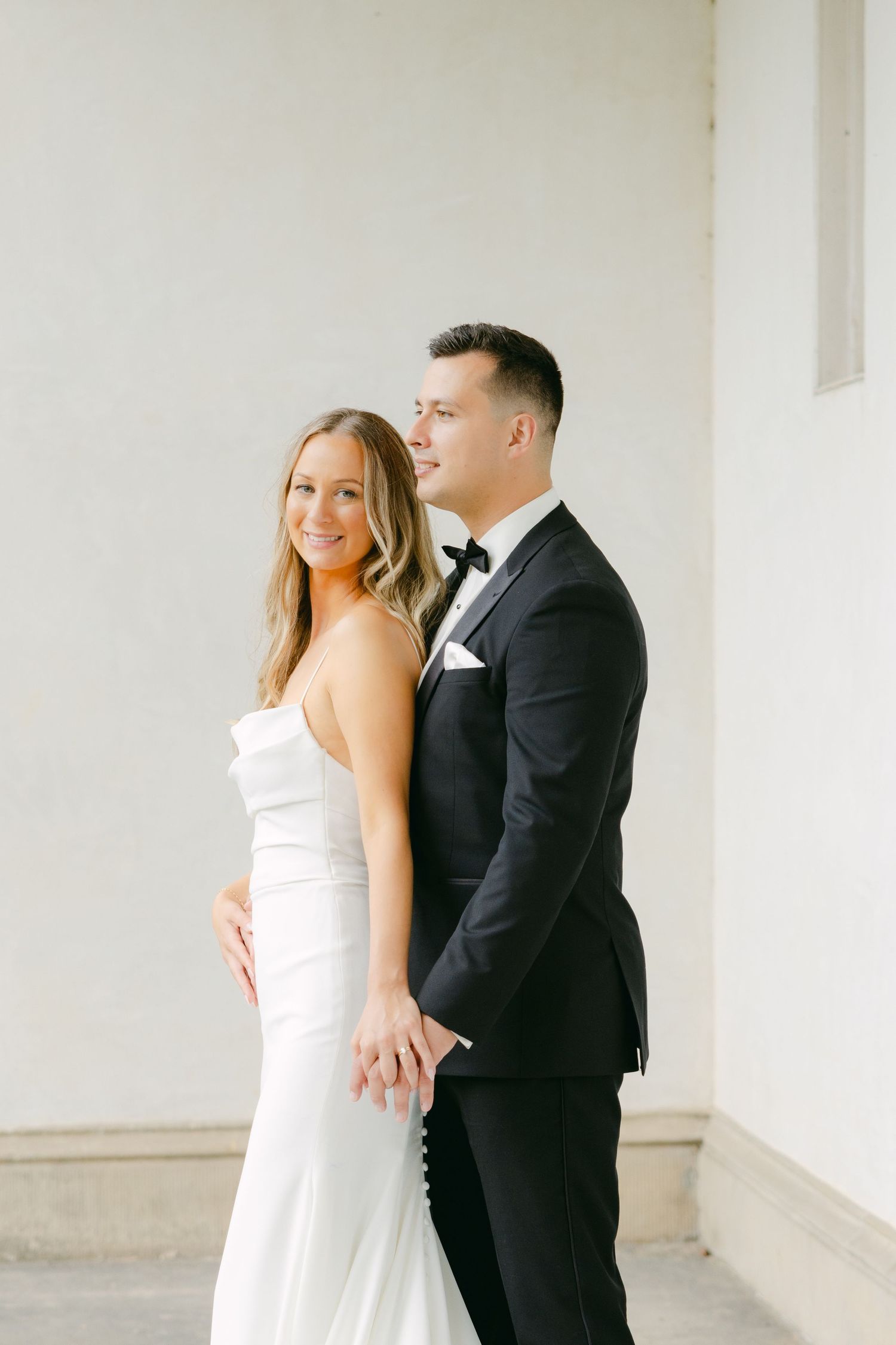 Elegant wedding couple posing together against white wall in formal attire.