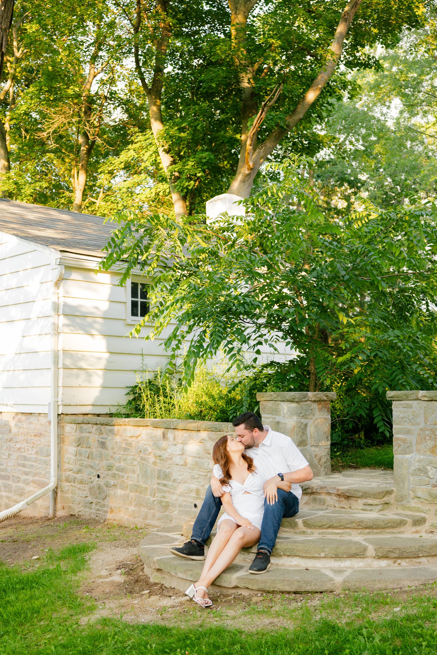 Summer engagement photo of couple on stone steps with white house and trees in background.