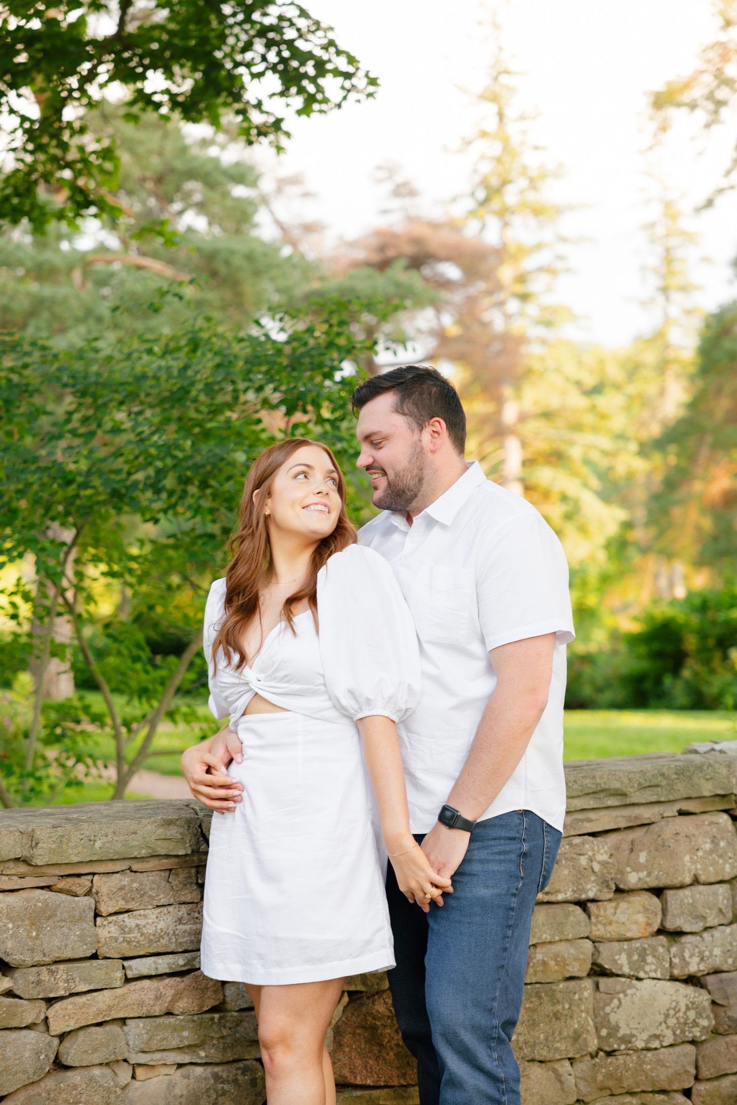 A couple leans against a stone wall in a garden setting during golden hour.