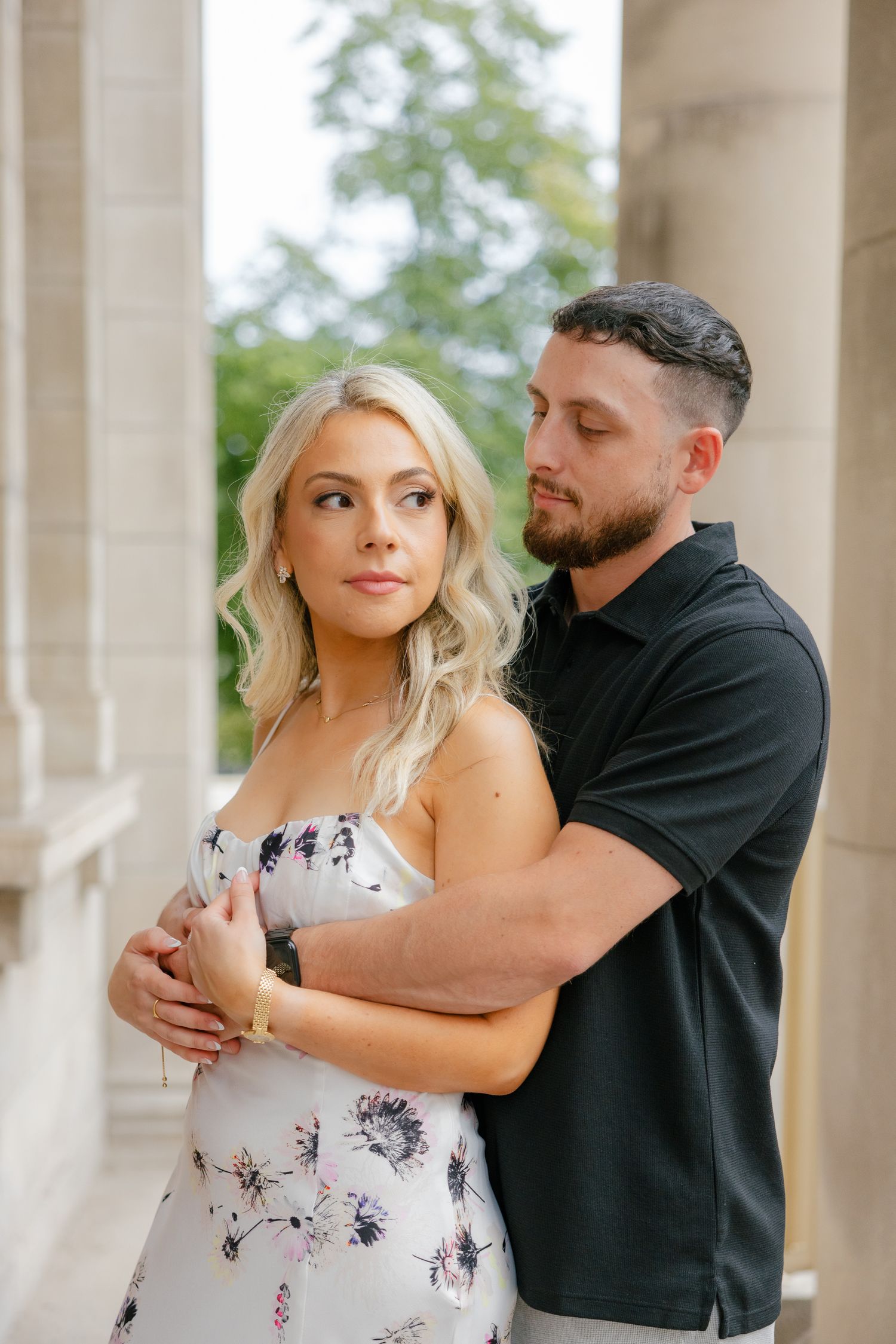 Couple in black polo and floral dress poses intimately between architectural columns.