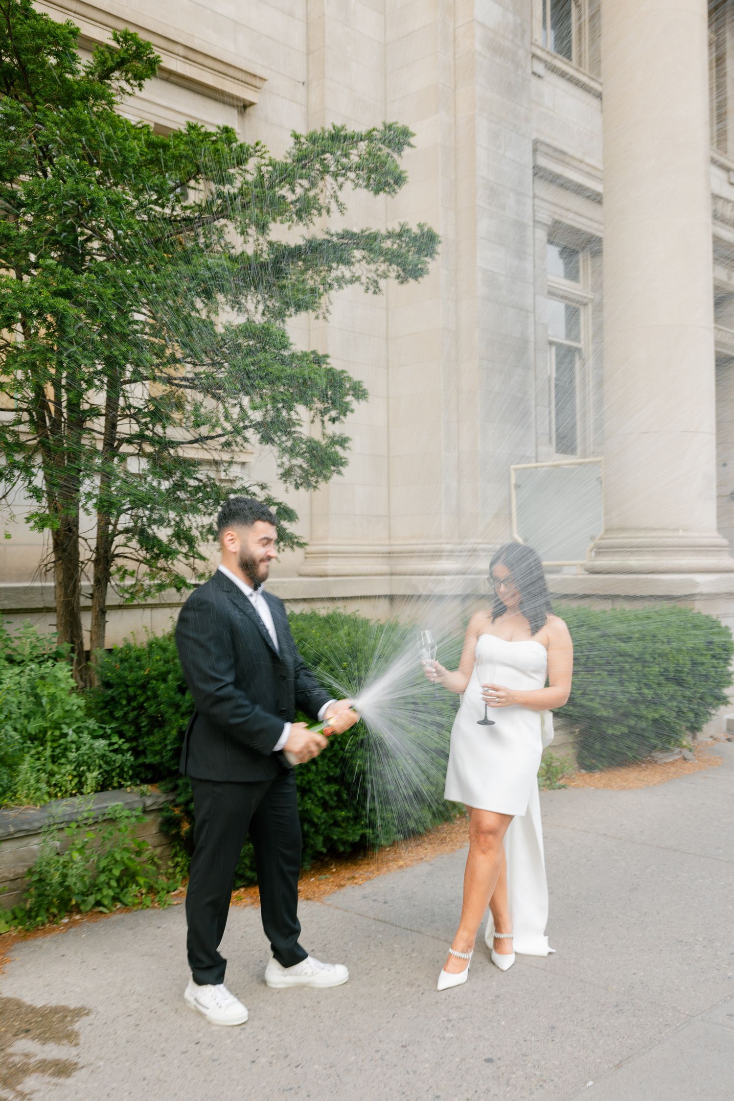 A champagne celebration moment captured outside a classic building with tree landscaping.
