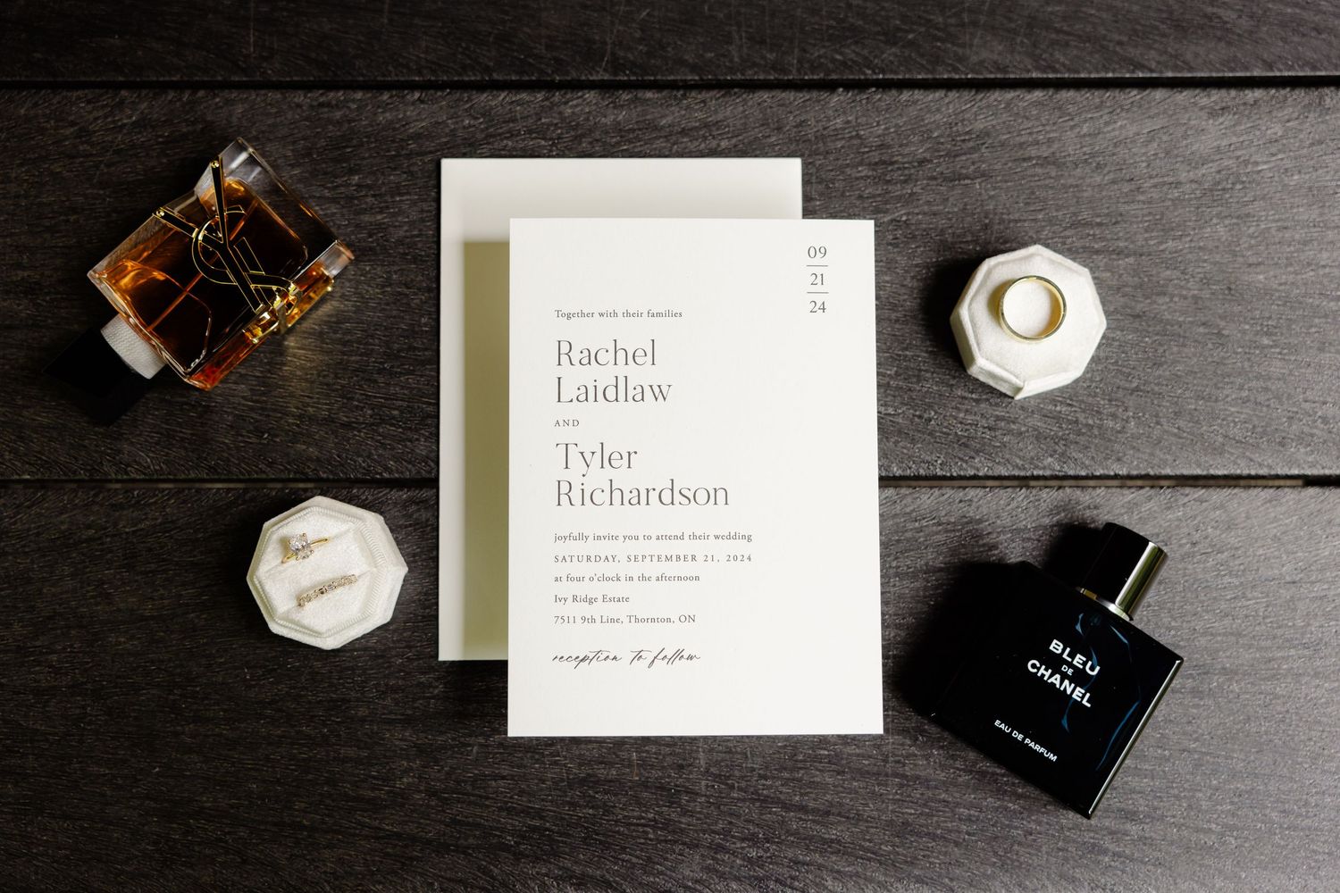 Wedding invitation flatlay with perfume bottles and ring boxes on dark wooden surface.