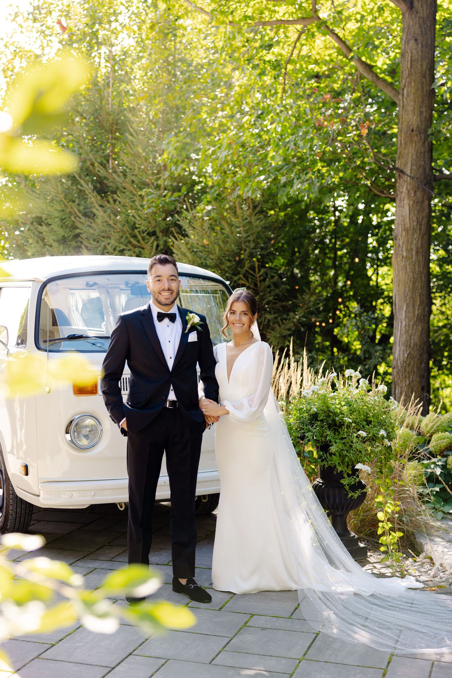 Wedding portrait featuring formal attire and a classic white vehicle against green garden scenery.