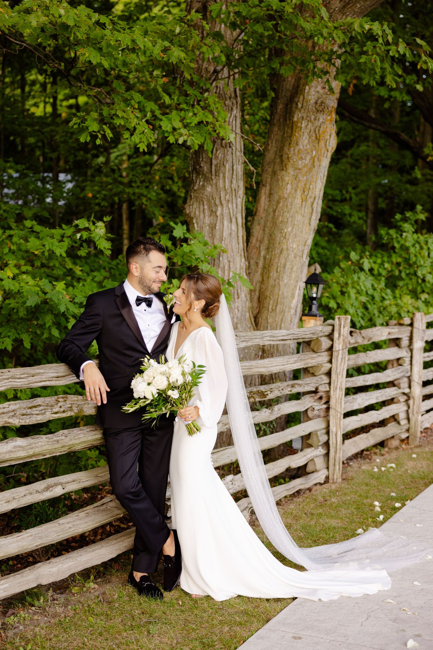 A romantic wedding photo against a rustic fence with the bride holding a white bouquet.