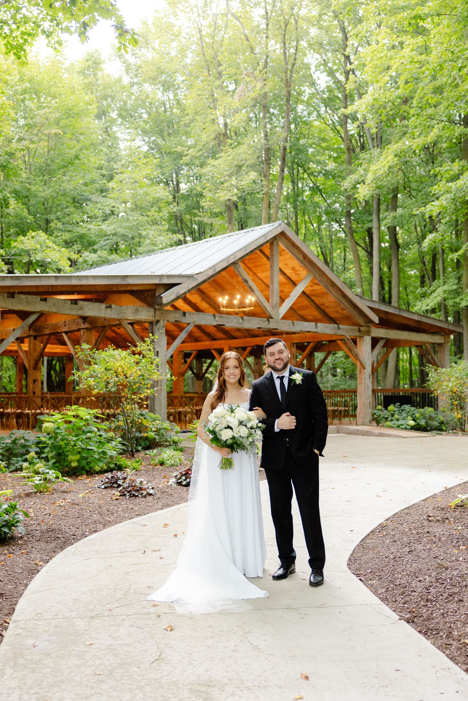 Couple walks along garden path with rustic timber pavilion and forest backdrop.