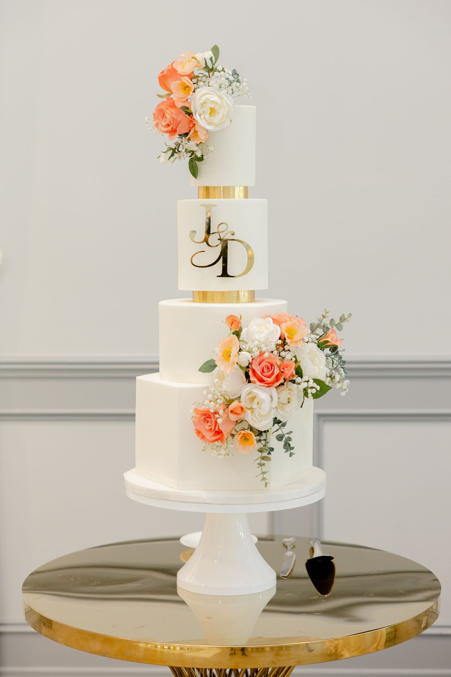 Four-tier white wedding cake adorned with coral and white roses and gold monogram details.