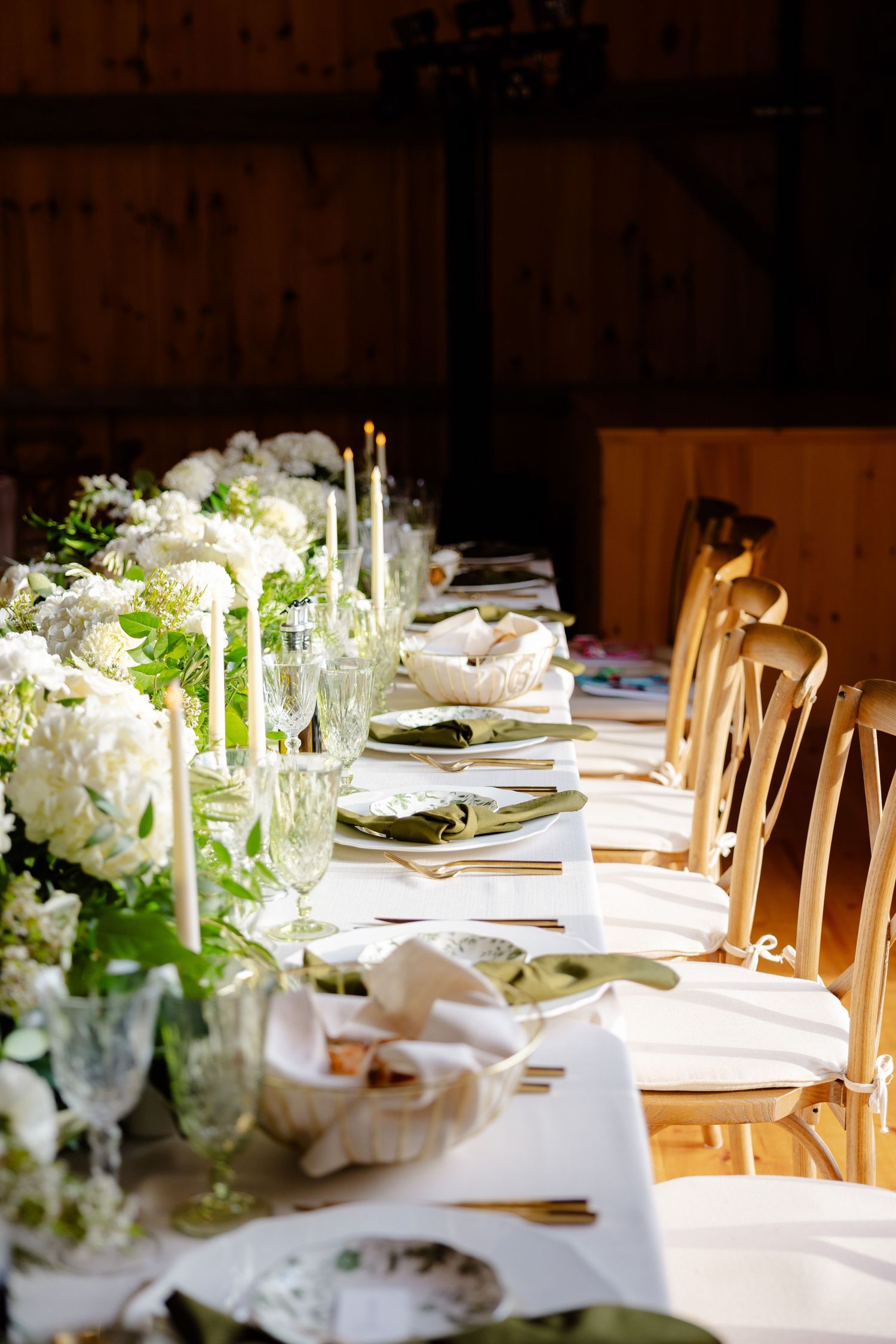An elegant wedding table setting with white floral centerpieces, taper candles, and gold chairs in a rustic venue.