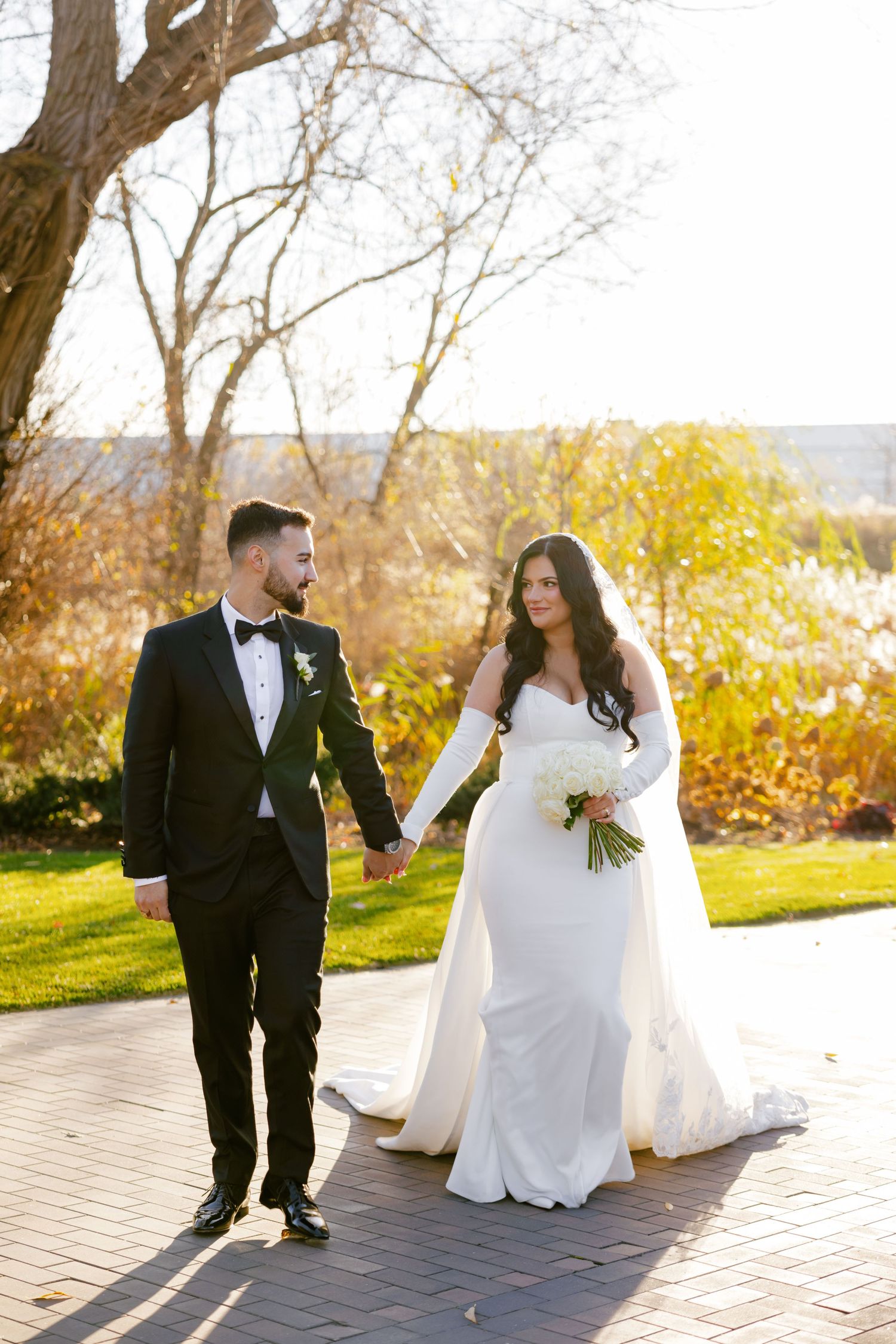 Bride in white gown and groom in tuxedo walking together on brick pathway during sunset.