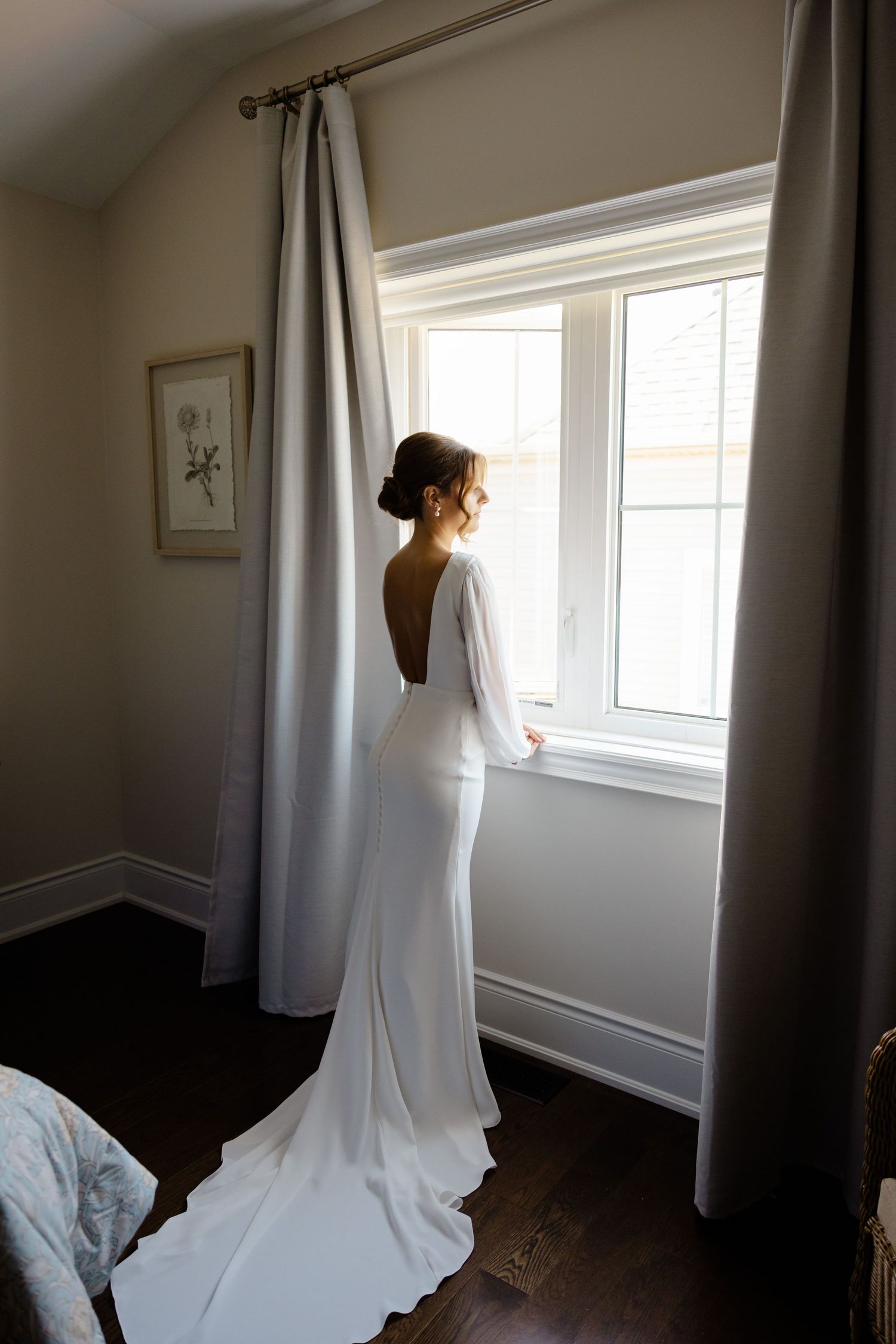 Backless wedding dress with flowing train photographed from behind near a bright window.