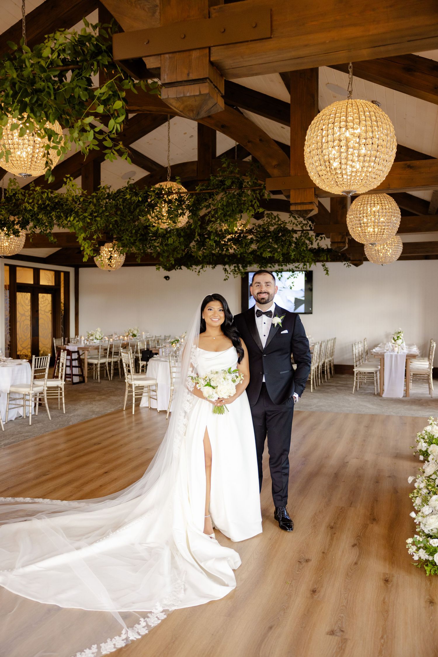 Elegant wedding venue with crystal chandeliers and greenery adorning wooden beams creates a romantic atmosphere.