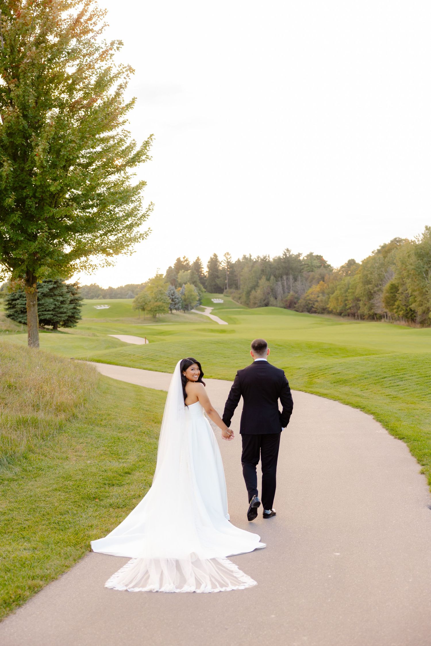 A newlywed couple walking on a golf course path with a long wedding dress train trailing behind.