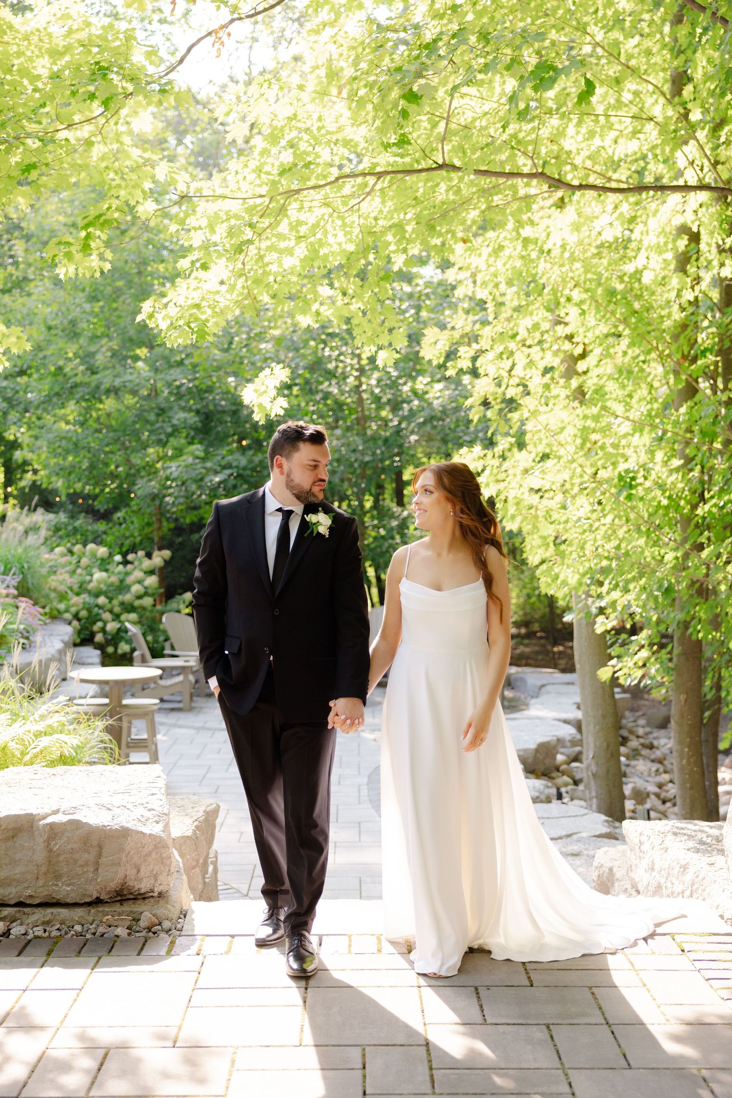 Newlyweds walk hand-in-hand along a stone pathway under sunlit tree branches in an elegant outdoor garden setting.