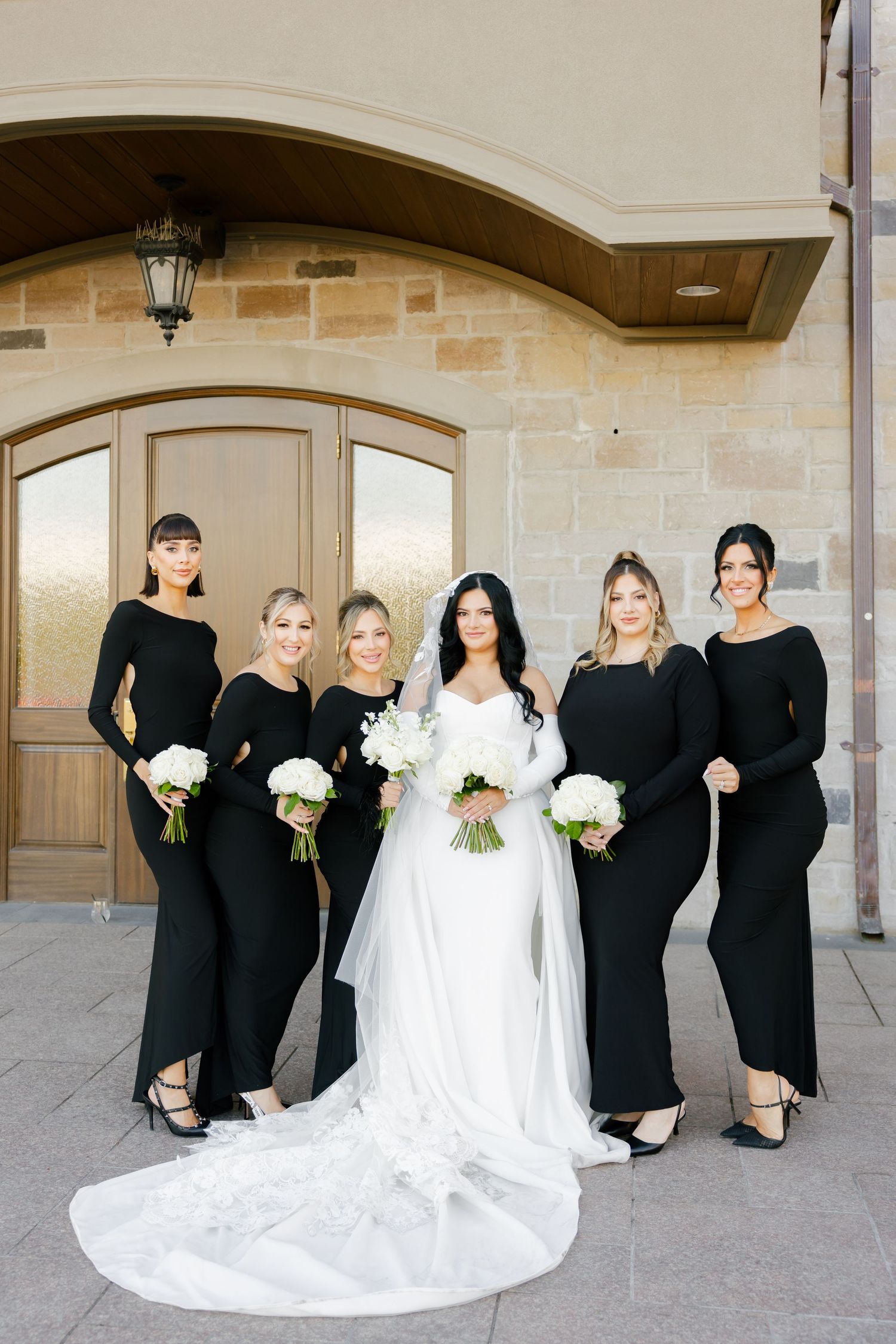 Bridal party poses in elegant black dresses and white bouquets against stone archway entrance.