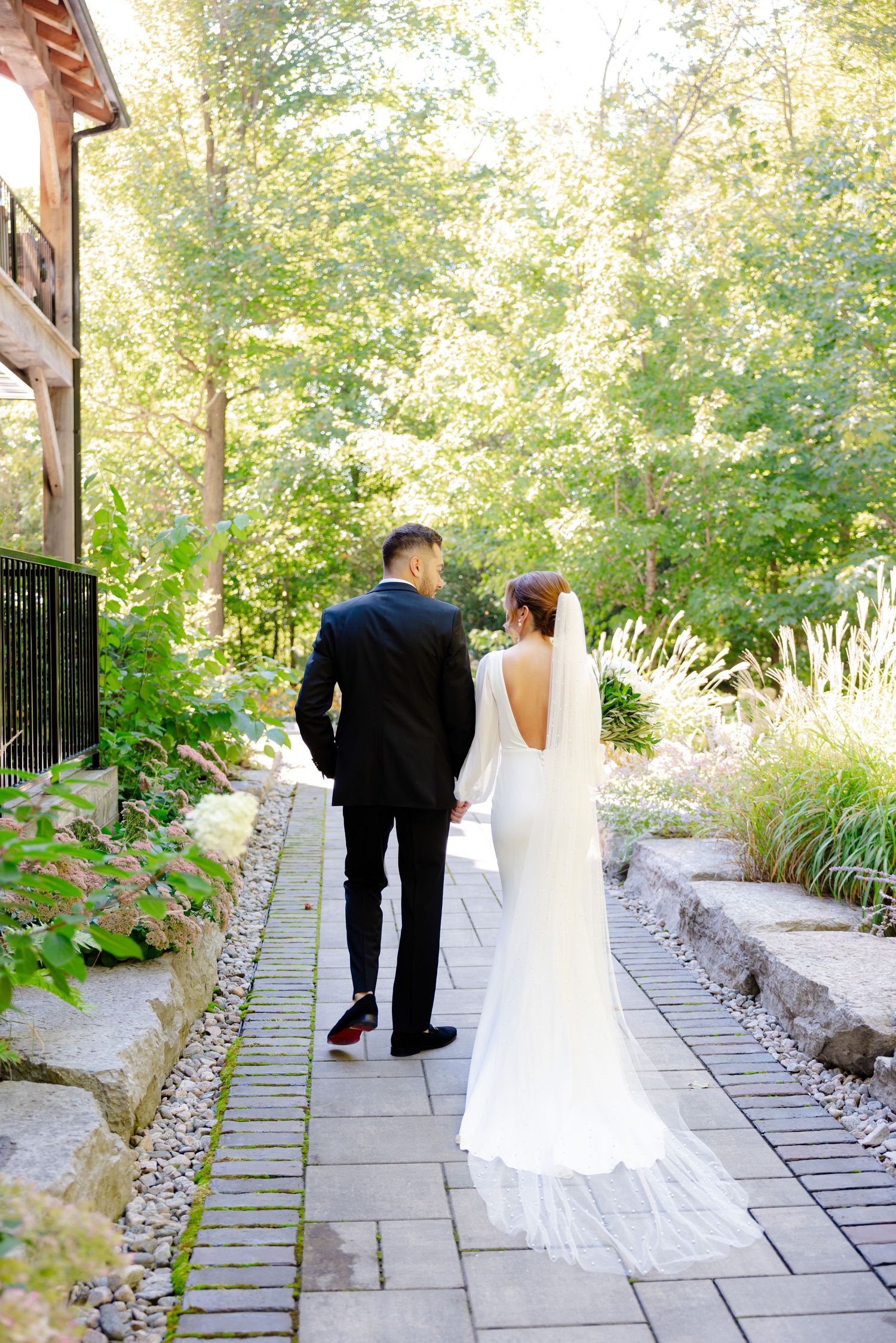 A couple in wedding attire walking along a stone garden path surrounded by lush greenery and trees.