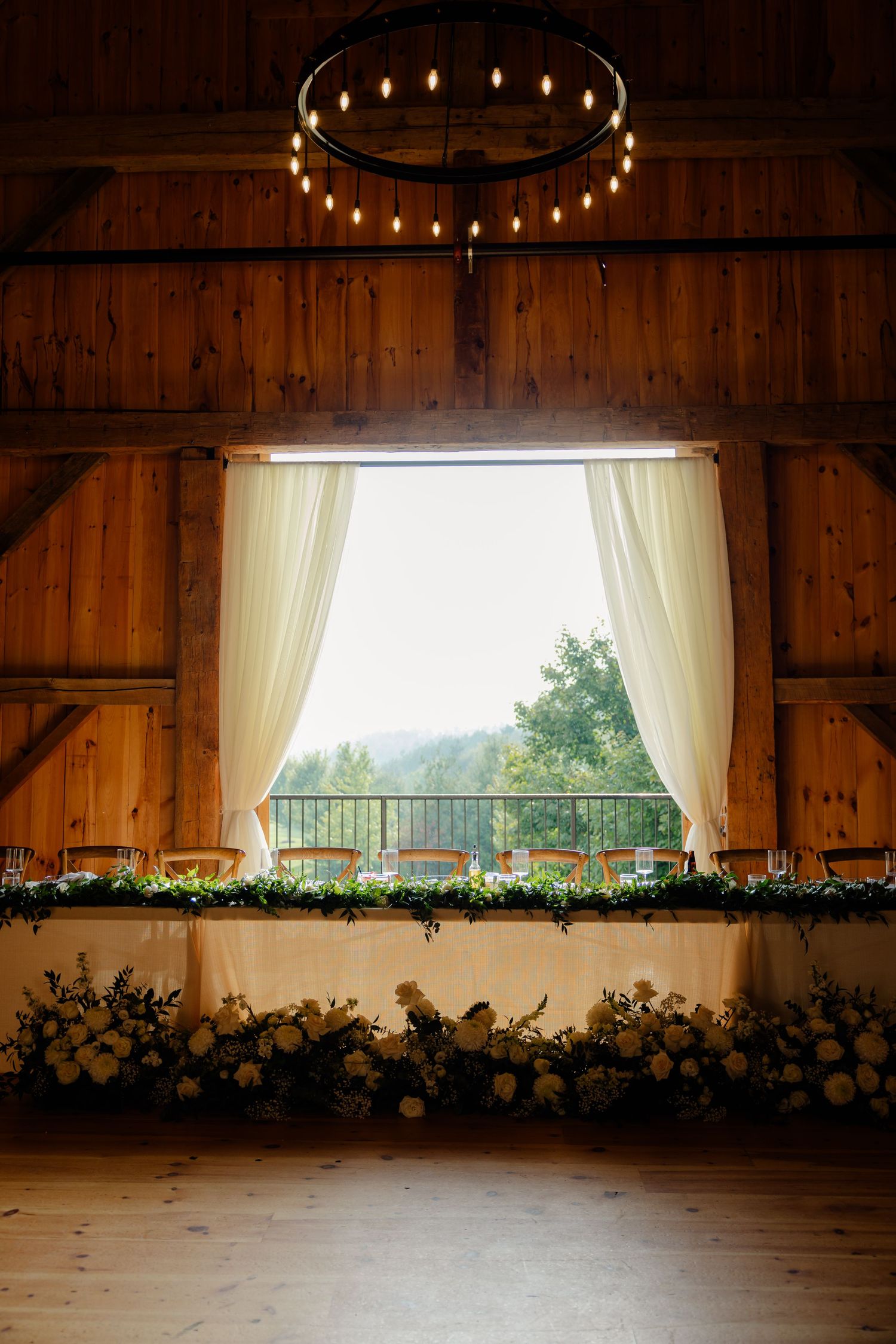 Barn wedding ceremony space with circular chandelier and flowing white curtains framing scenic window view.