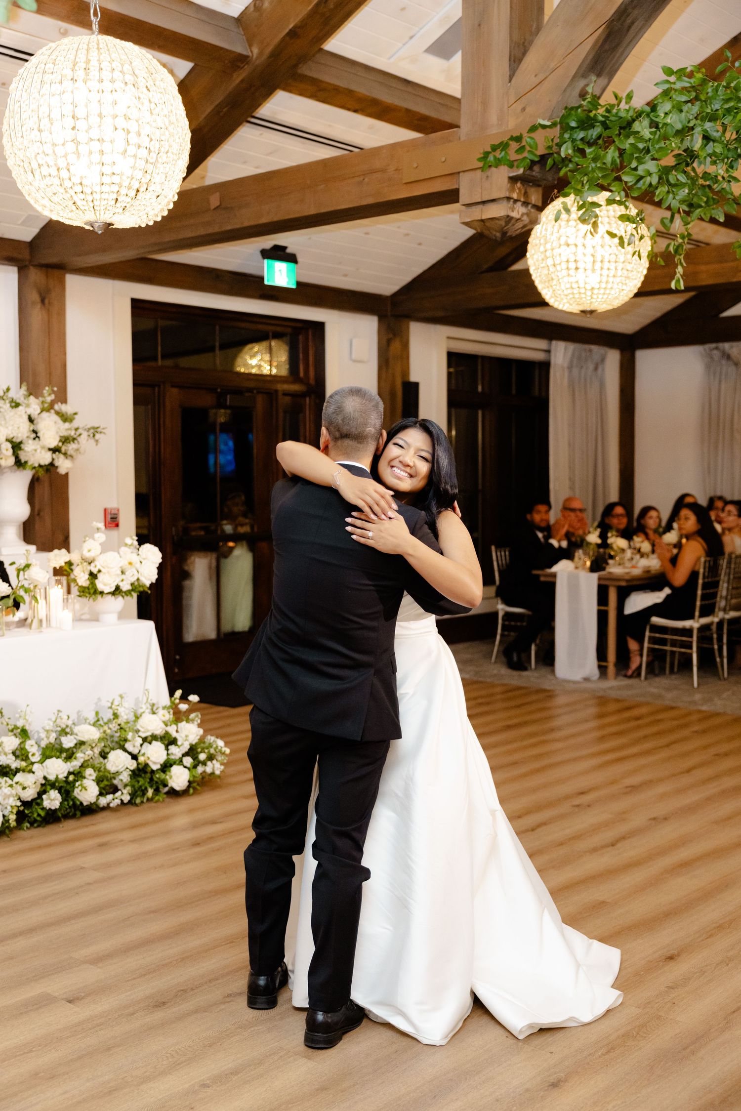 Wedding couple embraces during dance in beautiful hall with crystal chandeliers and wooden beams.