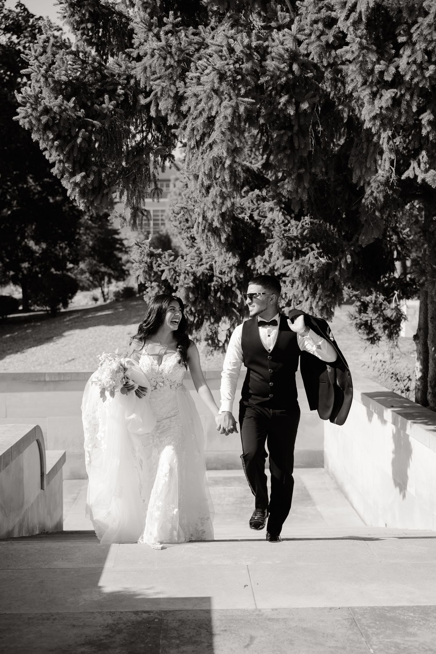 A black and white wedding photo of a couple walking up stairs under overhanging trees.