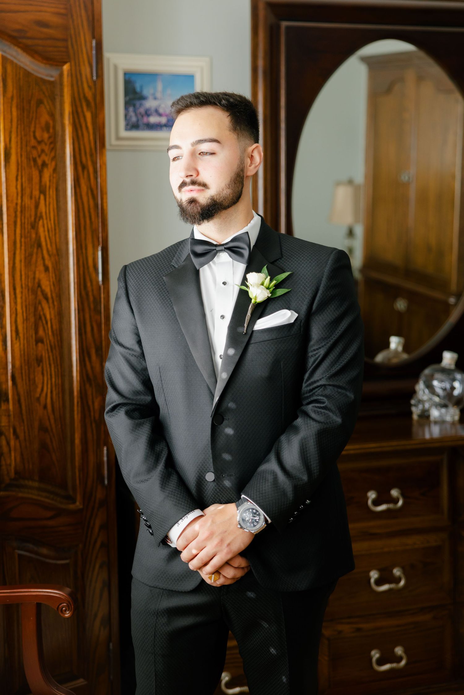 A groom wearing a black tuxedo and boutonniere stands confidently with clasped hands.