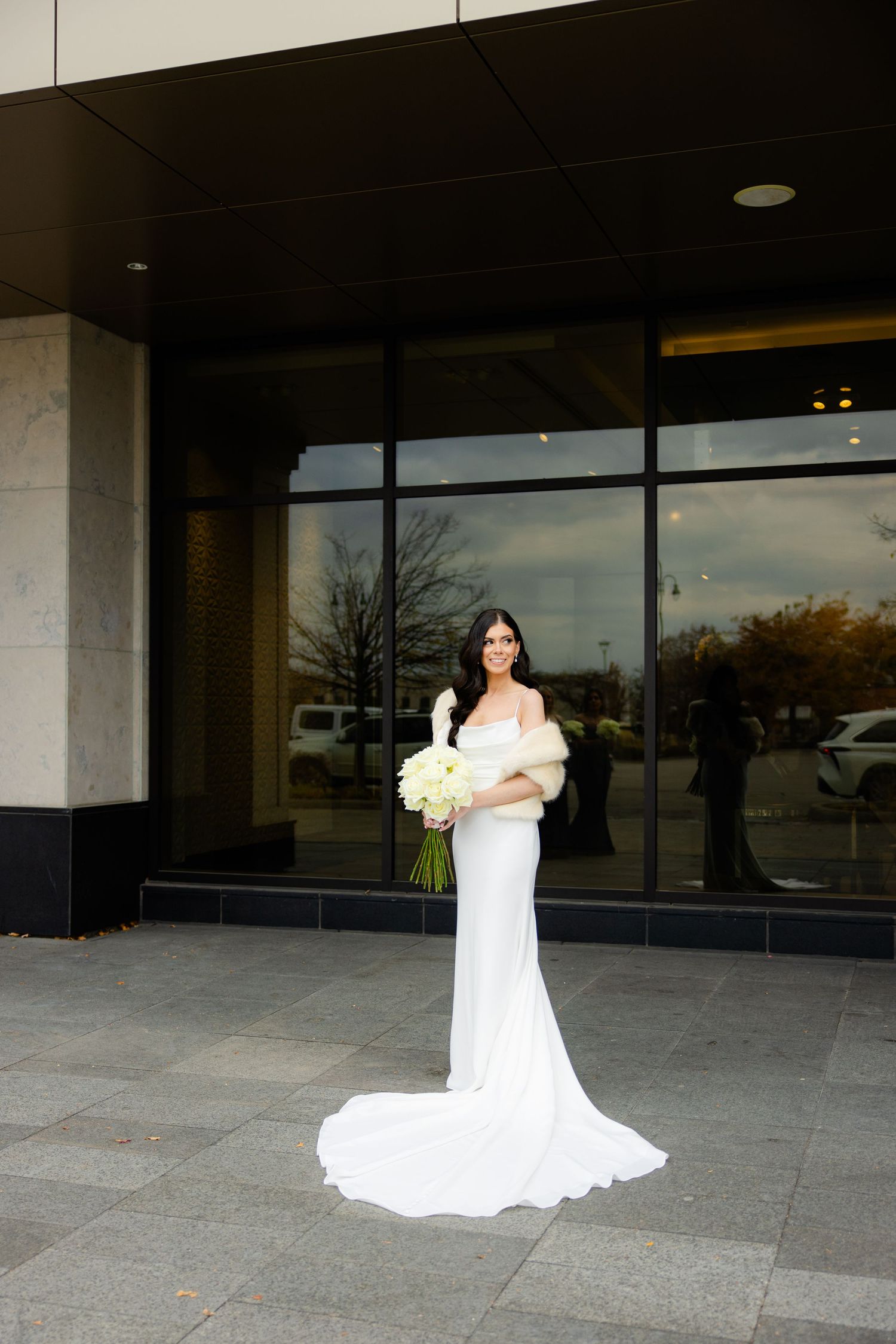 Elegant bride in fitted white gown holds cream bouquet while standing against modern building exterior.