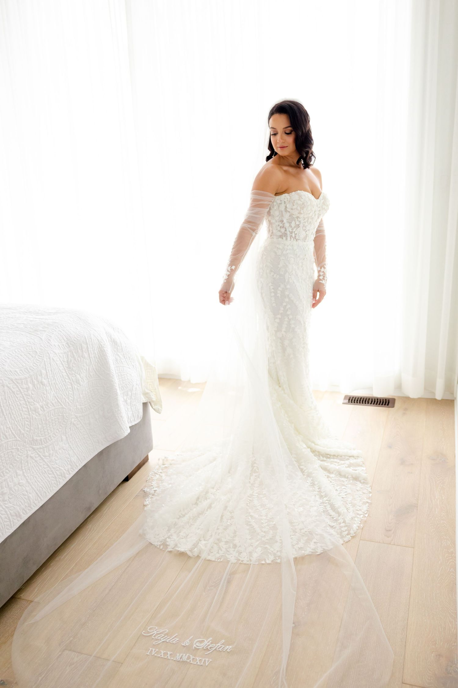 A detailed shot of a fitted lace wedding dress with off-shoulder design and cathedral train on hardwood floors.