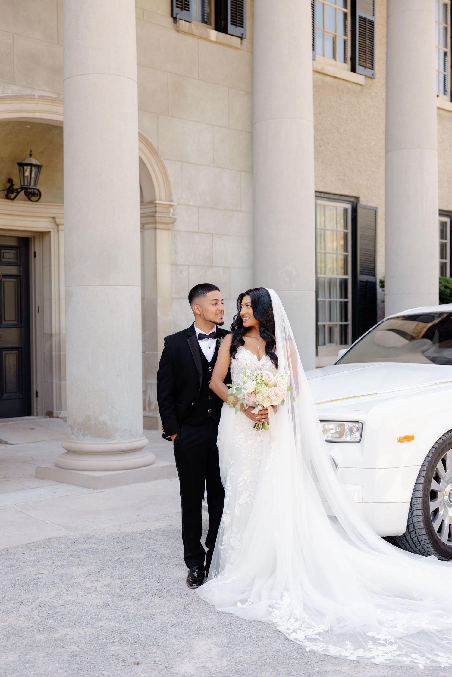 Romantic wedding photo with couple standing beside white luxury car and stone pillars.